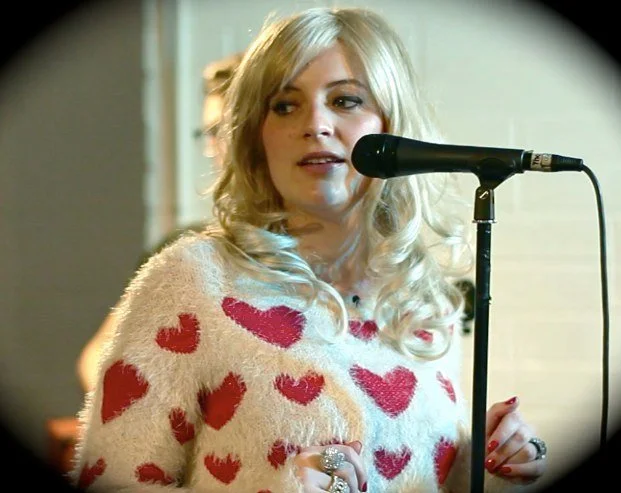 A blonde woman wearing a white sweater with red hearts, speaking into a black microphone.