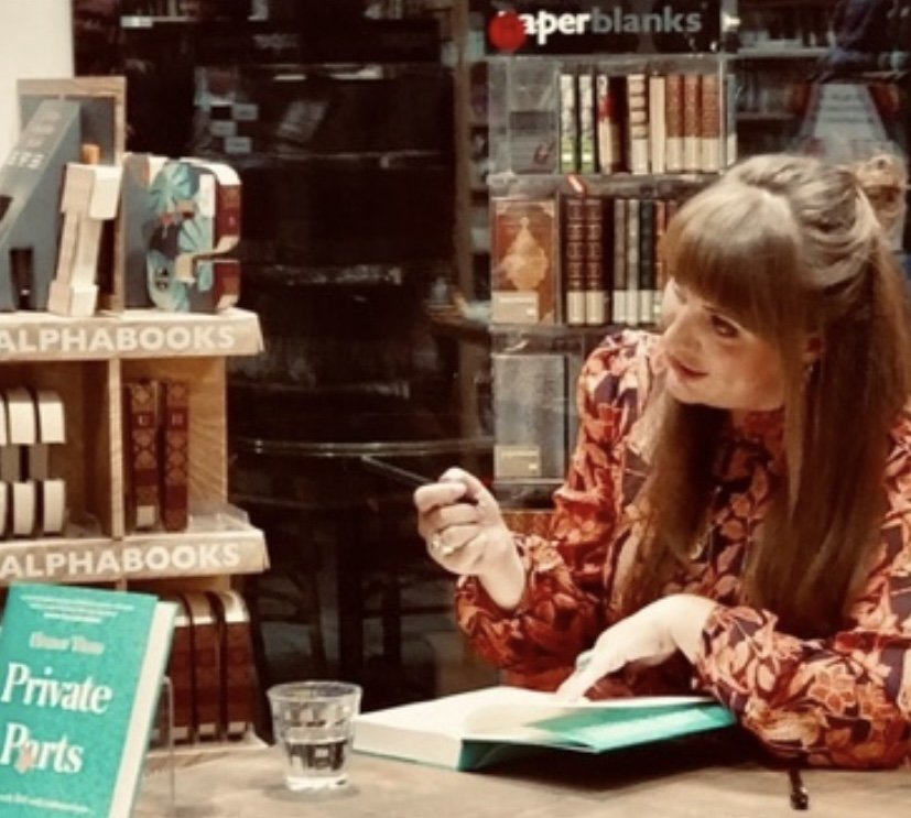 A woman with long brown hair tied back, wearing a floral patterned shirt, is sitting at a table in a bookstore. She is holding a pen and is pointing to an open book. There is a glass of water on the table and shelves of books behind her, including a bookshelf labeled 'ALPHABOOKS' and another for 'superblanks'.