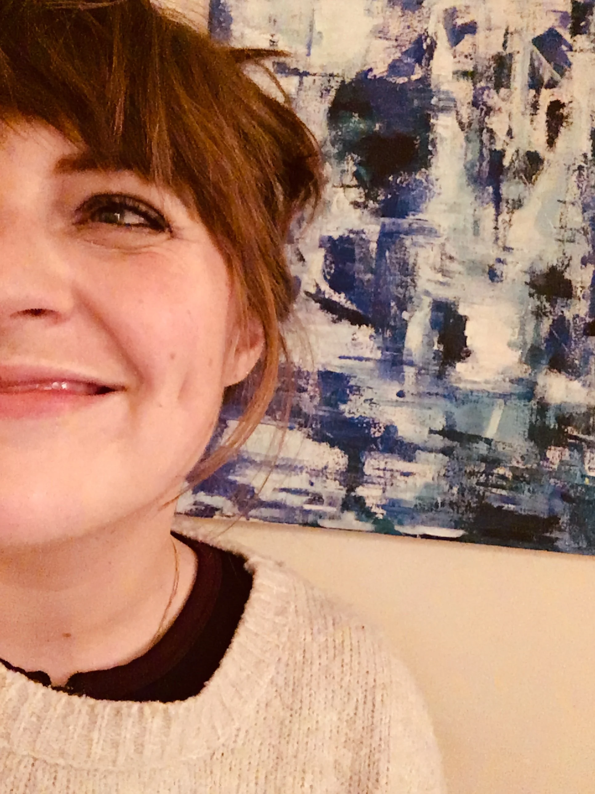 Partial close-up of a woman with brown hair, smiling, wearing a cream-colored sweater, standing in front of a blue and white abstract painting.