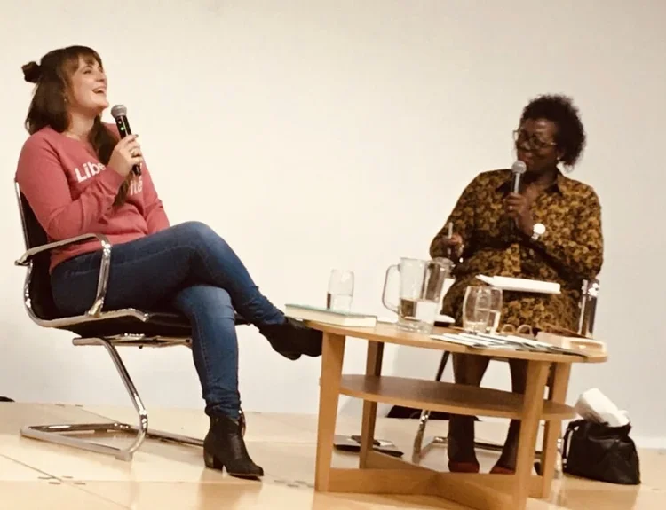 Two women sit on stage during a discussion, each holding a microphone. One woman is wearing a pink hoodie and jeans, smiling, while the other is wearing a patterned blouse, listening attentively, an event at Sheffield book festival 'Off The Shelf'.