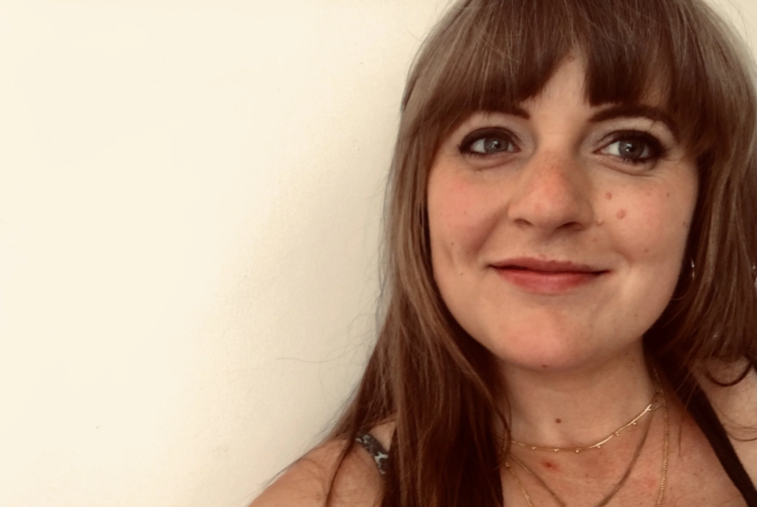Close-up of a woman with light skin, reddish-brown hair, blue eyes, and freckles, wearing a black top with patterned straps and layered necklaces, smiling softly against a plain, light-colored wall.