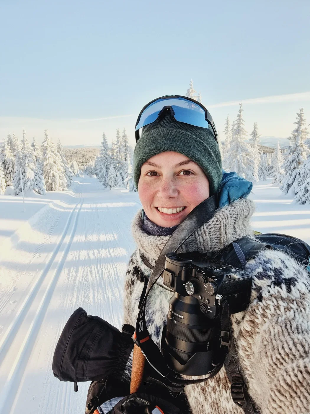 A selfie of a woman in a woolen sweater and hat smiling into the camera. In the background is a snow covered landscape and cross-country skiing tracks