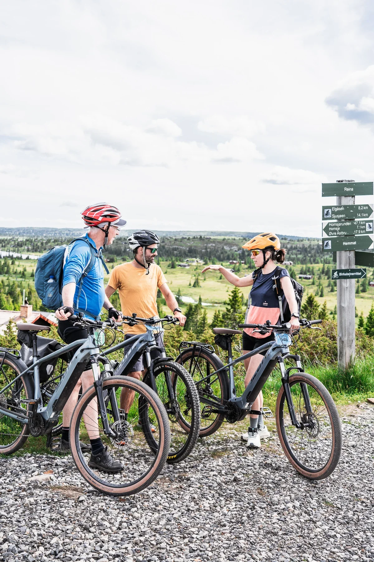 A group of 3 cyclists standing next to their gravel bikes and talking with each other. All three wear helmets and small backpacks. In the background is a signpost and a wide open landscape