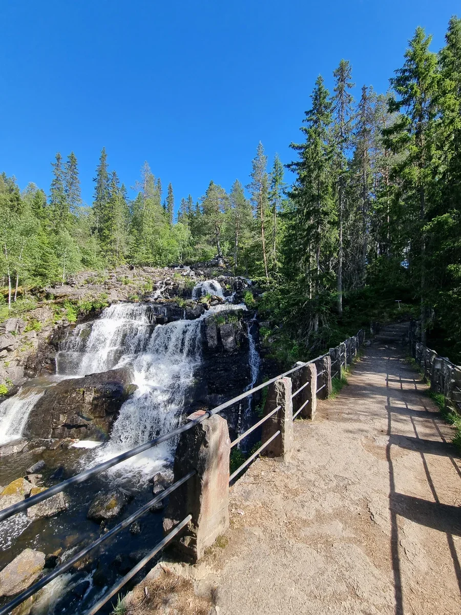 A path with fences to both sides leads along a waterfall cascading downhill over the rocks. All around you can see green trees.