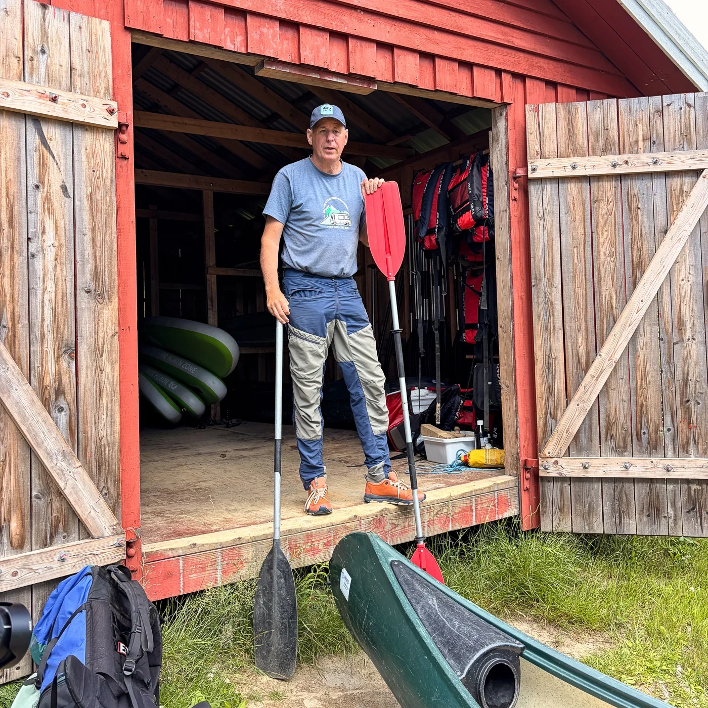 A man in outdoor clothing and a tshirt standing in the door to a shed holding two paddles. In the foreground the tip of a green canoe is visible.