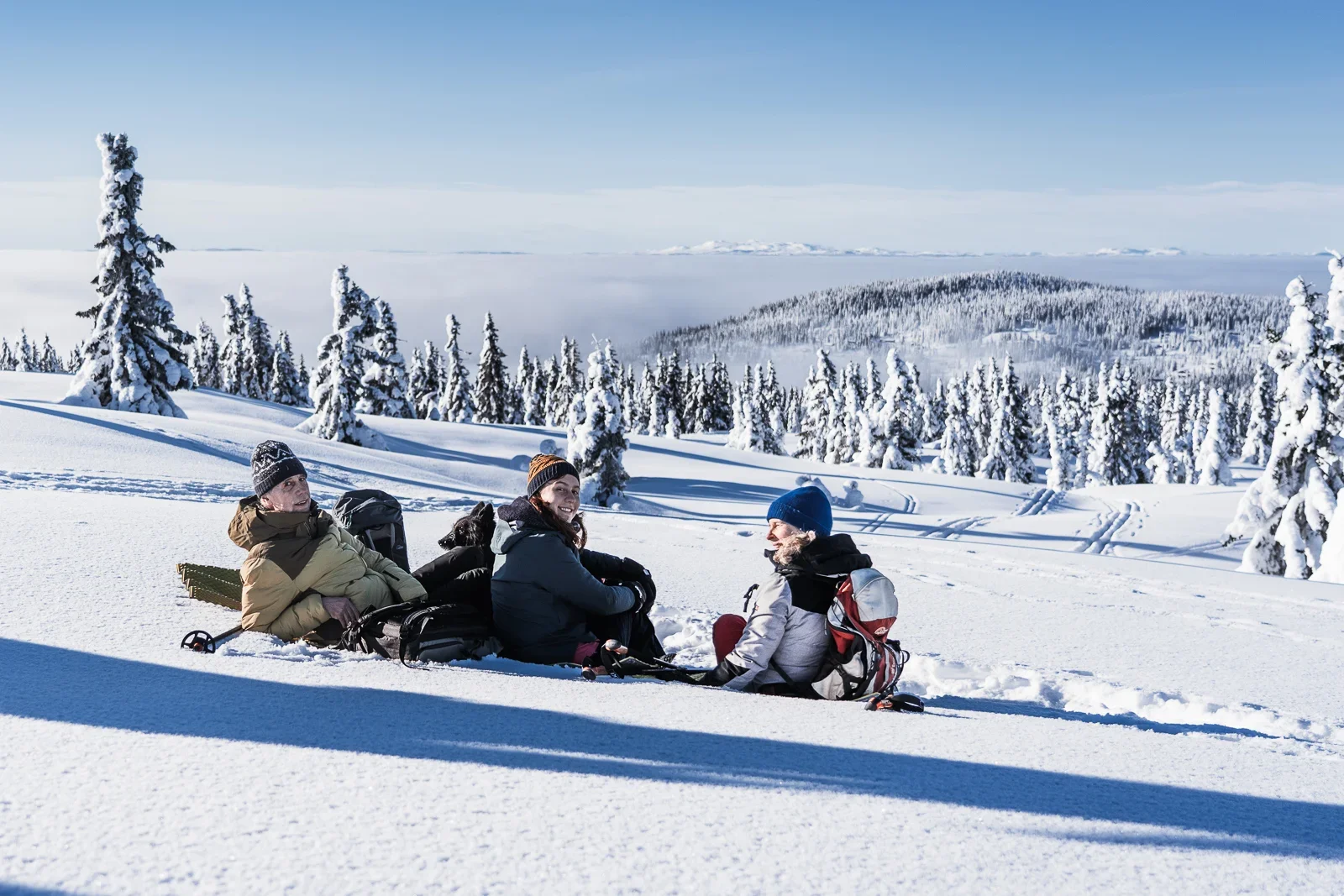 A family of three and a dog sitting in the snow in front of a wide hilly winter landscape. In the background you can see snow-covered trees and a hill sticking out of the fog in the valley.
