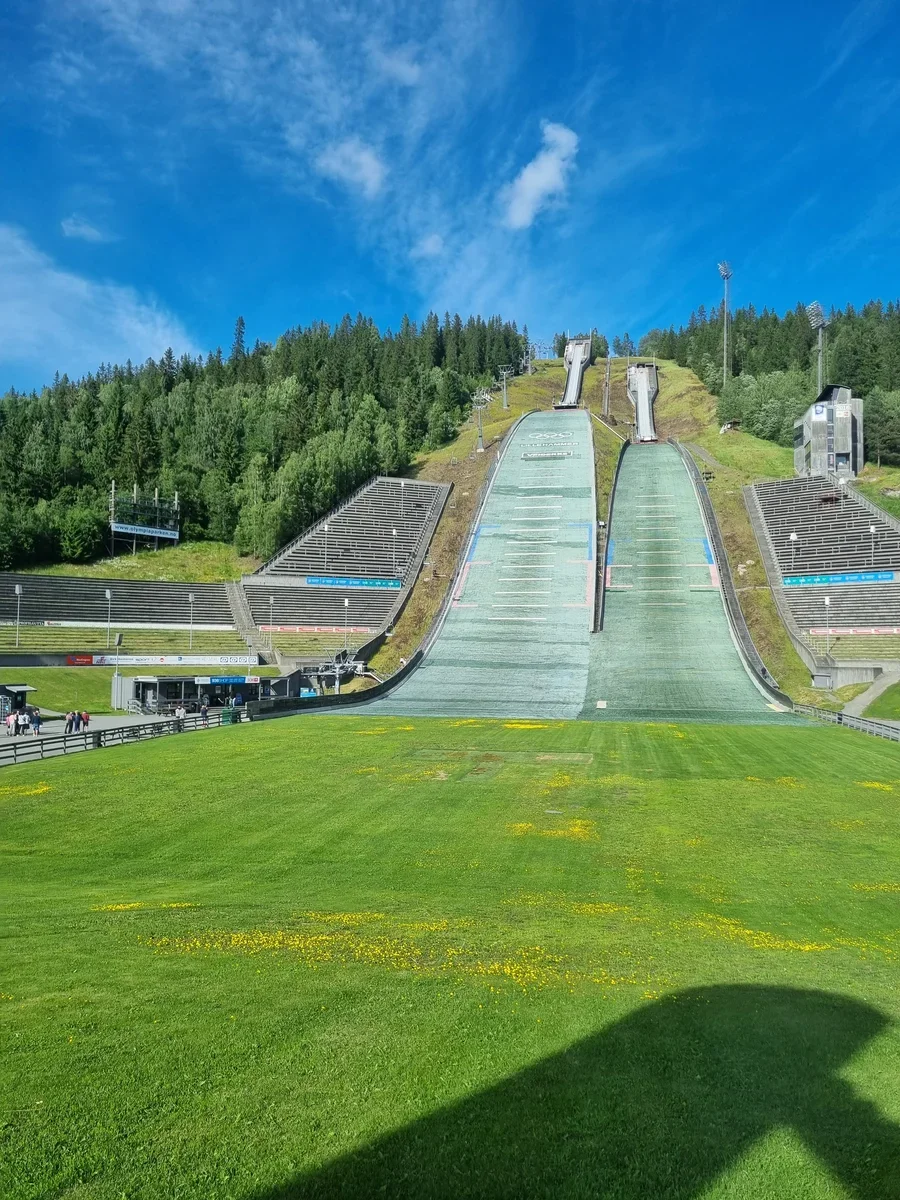 A photo from the bottom of two ski jumps in summer in Lillehammer. On both sides of the jump are empty stands.