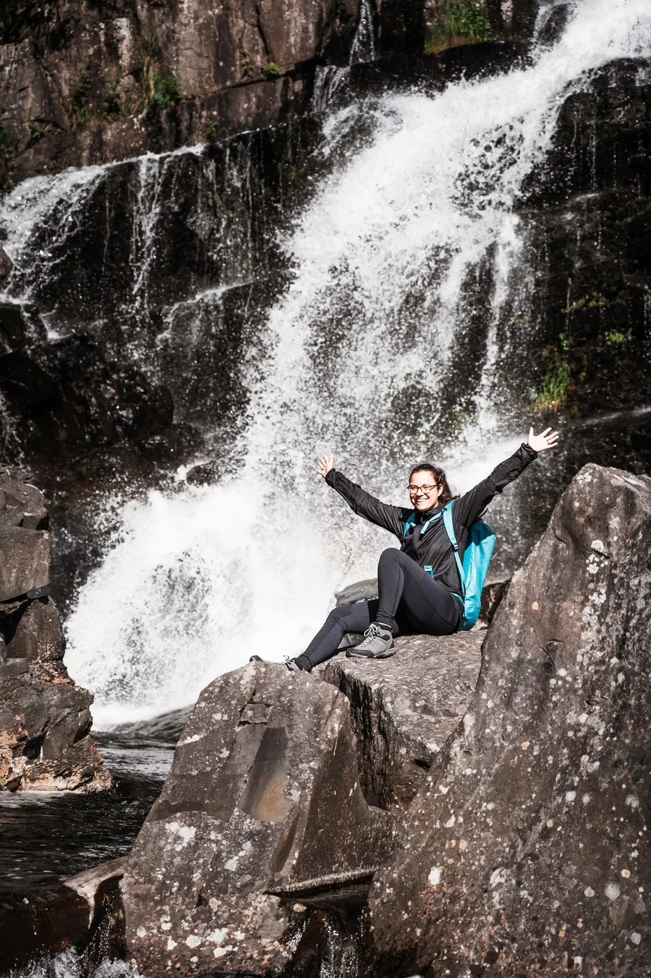 A woman sitting on some rocks in front of a gushing waterfall