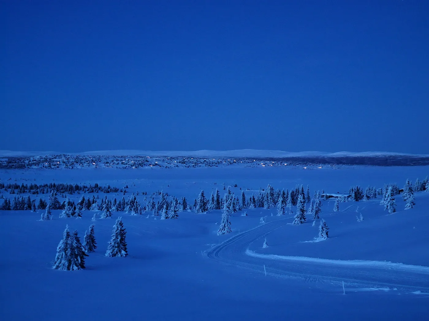 A wintry landscape in the dark. At the horizon a few lights of cabins are visible