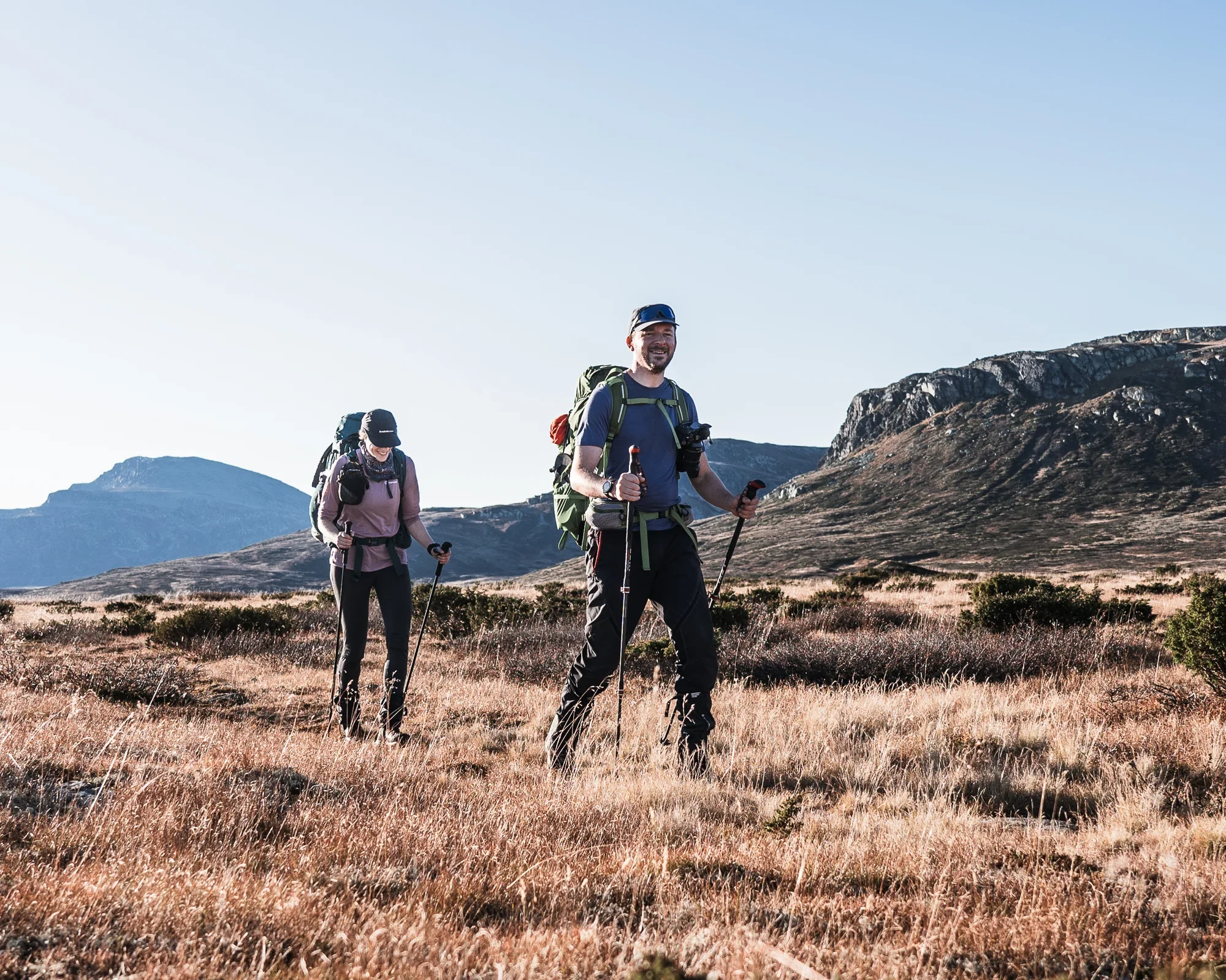 Two smiling people hiking through a grassy landscape with mountains in the background