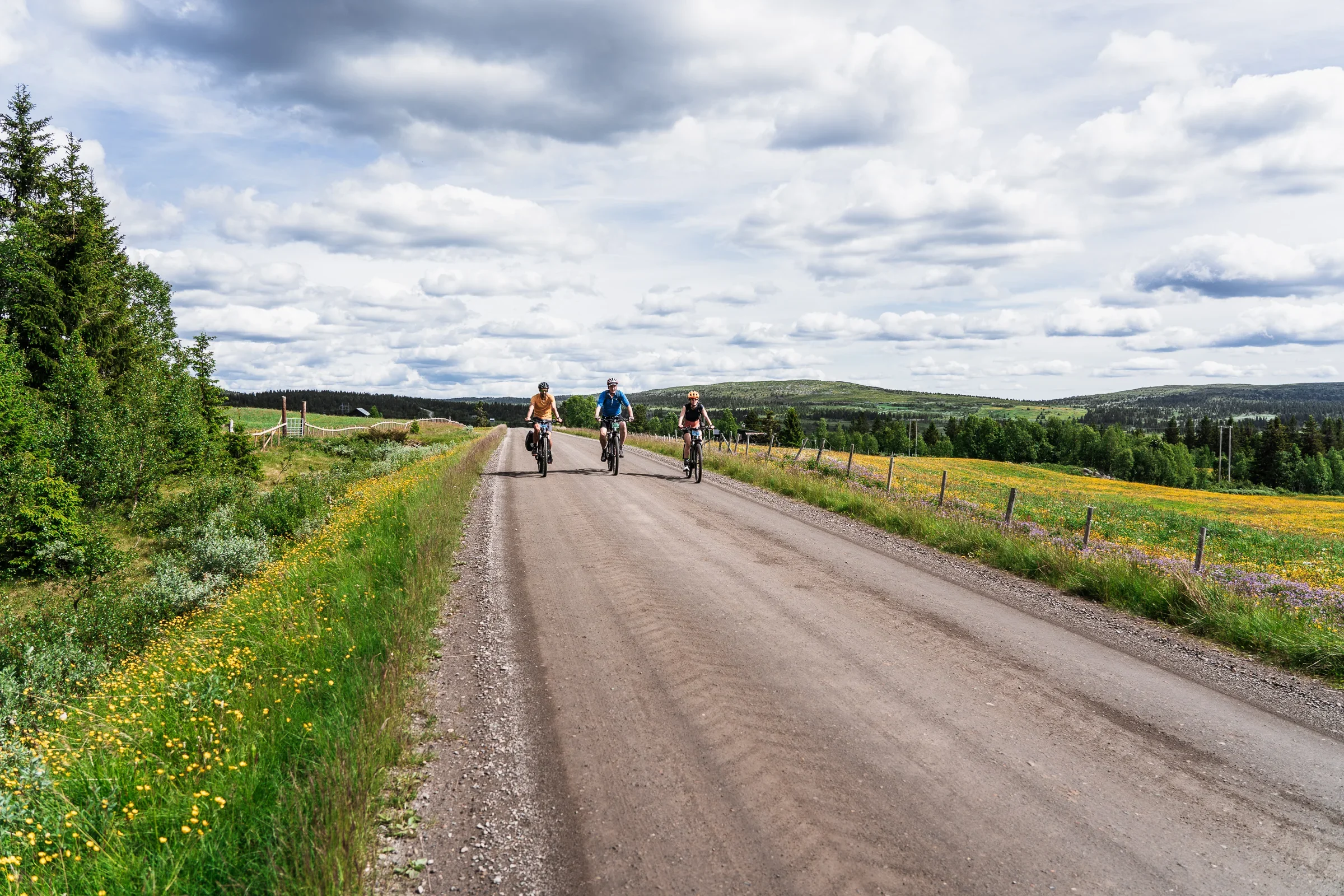 Three cyclists cycling next to each other towards the camera on a gravel road. In the background you can see green hills and a cloudy sky