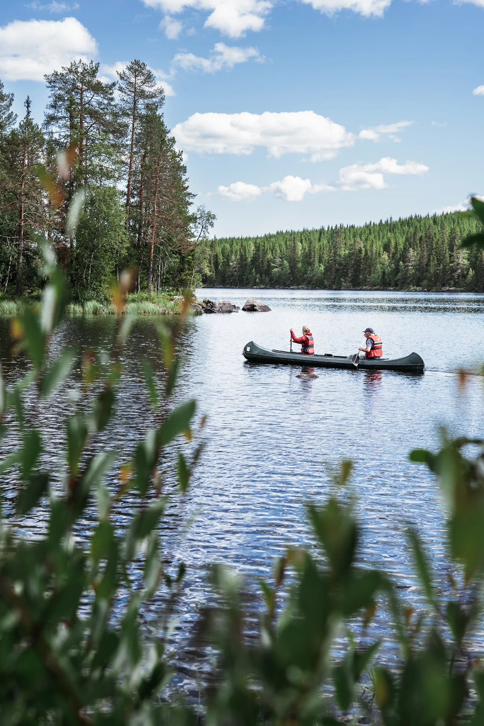 A couple sitting in a canoe paddling over a lake. In the foreground blurry bushes are visible. In the background you can see the shoreline and islands with trees.