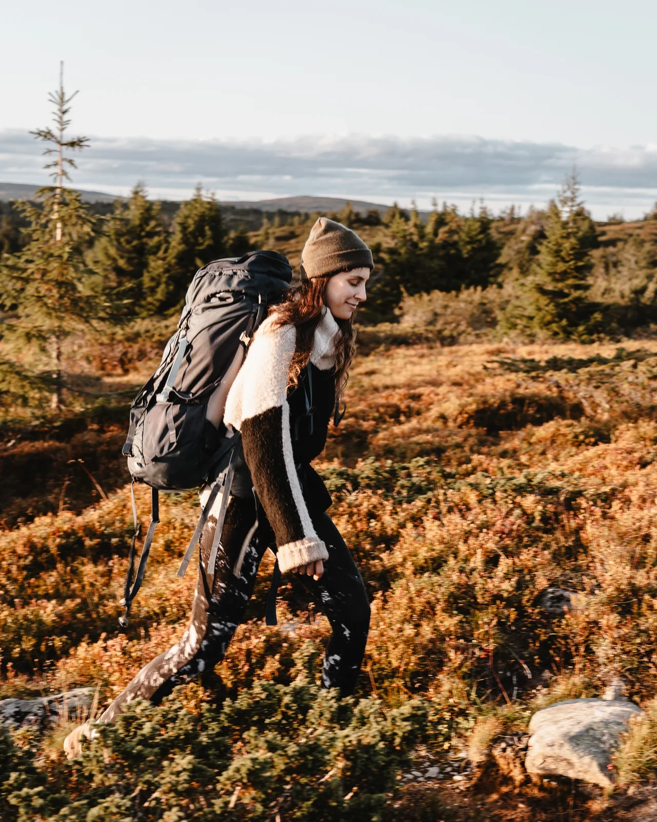 A woman with a wool sweater and hat and a big backpack hiking. In the background you can see some fir trees and rolling hills covered in warm sunlight