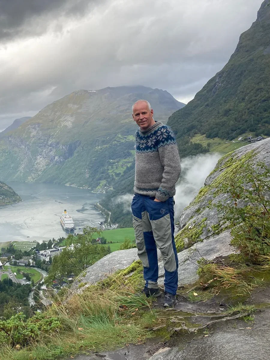 A photo of a man with woolen sweater and hiking pants standing on a mountain. In the background you can see a Norwegian fjord.