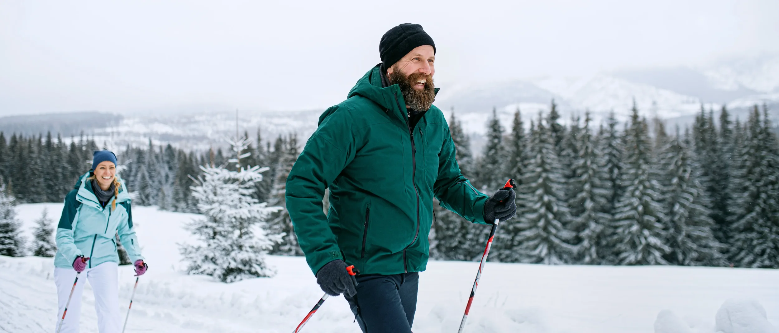 A man with a beard and a young woman wearing winter clothes and skiing on cross-country skis. In the background you can see a snow covered winter landscape.