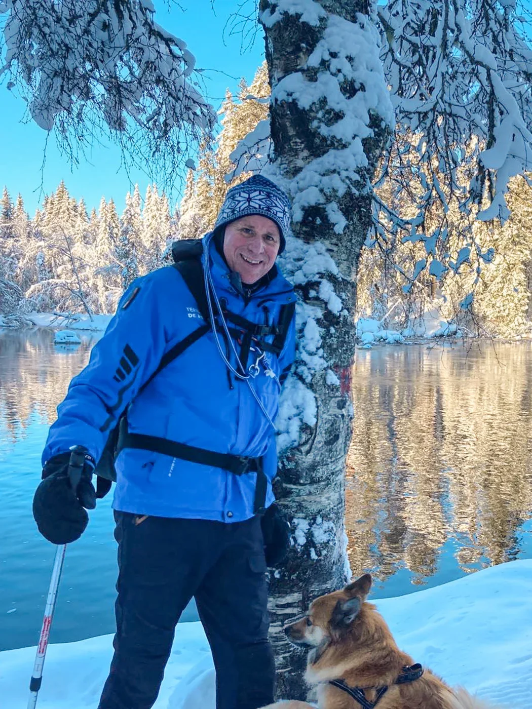 A photo of a man in winter clothing standing in front of a snow covered birch tree and a lake