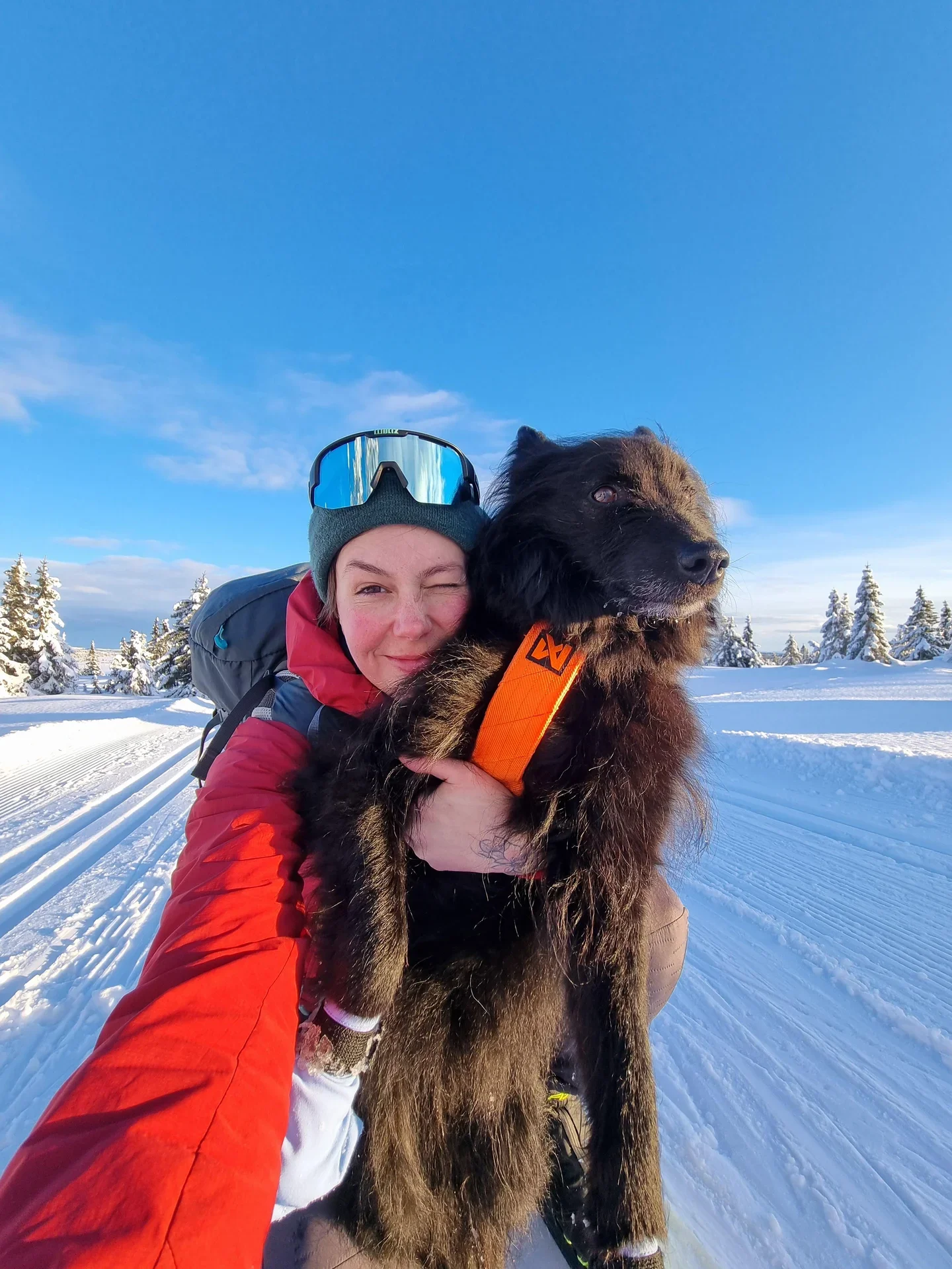 A selfie of a woman in a red jacket holding her dog and looking into the camera with a ski track and wintry landscape in the background