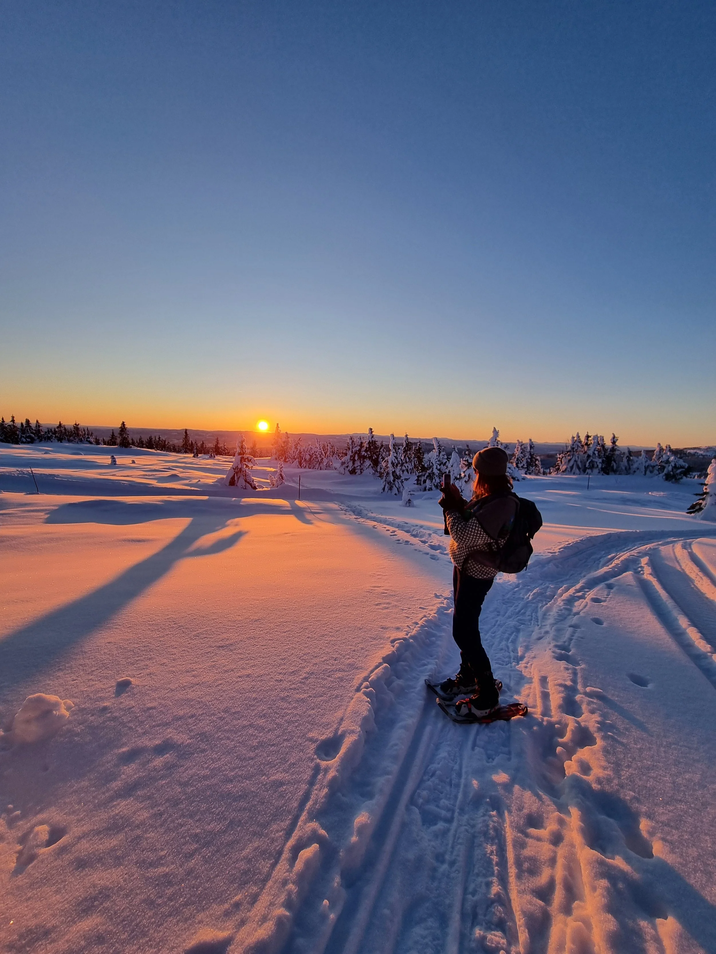 A woman on snowshoes standing in a wintry landscape taking a photo of the sunset