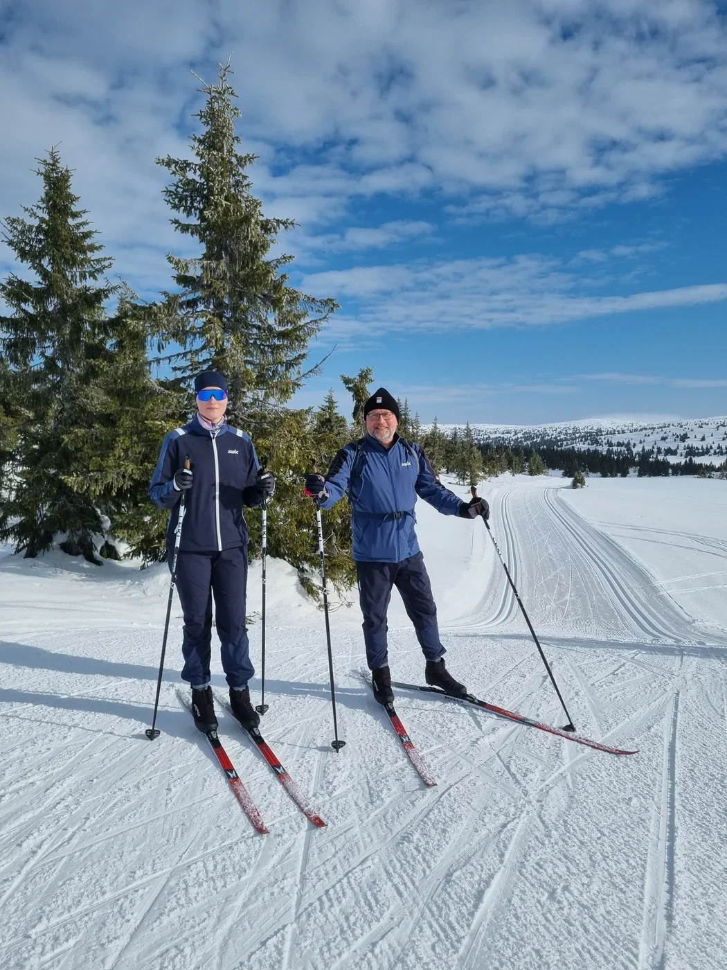Two people on cross-country skis standing in front of a wide winter landscape, smiling into the camera