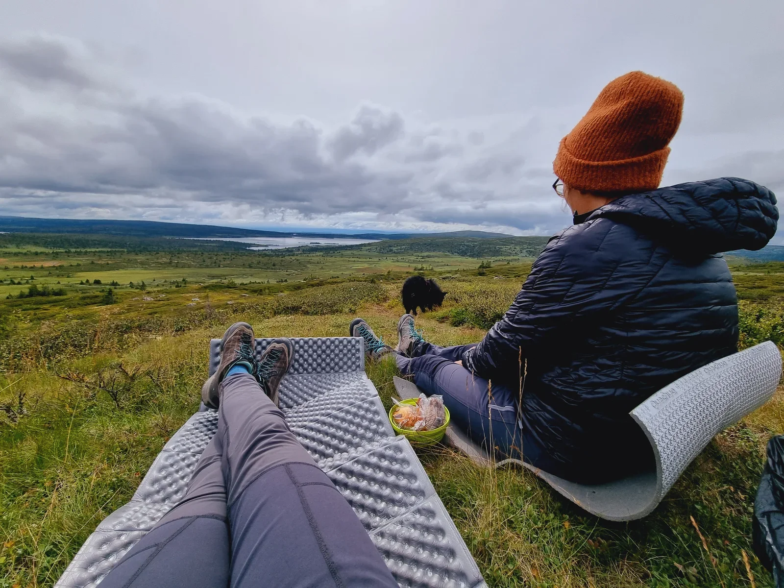 Two hikers sitting on foam mats enjoying a break. You can see the legs of one person and a second person sitting next to it.