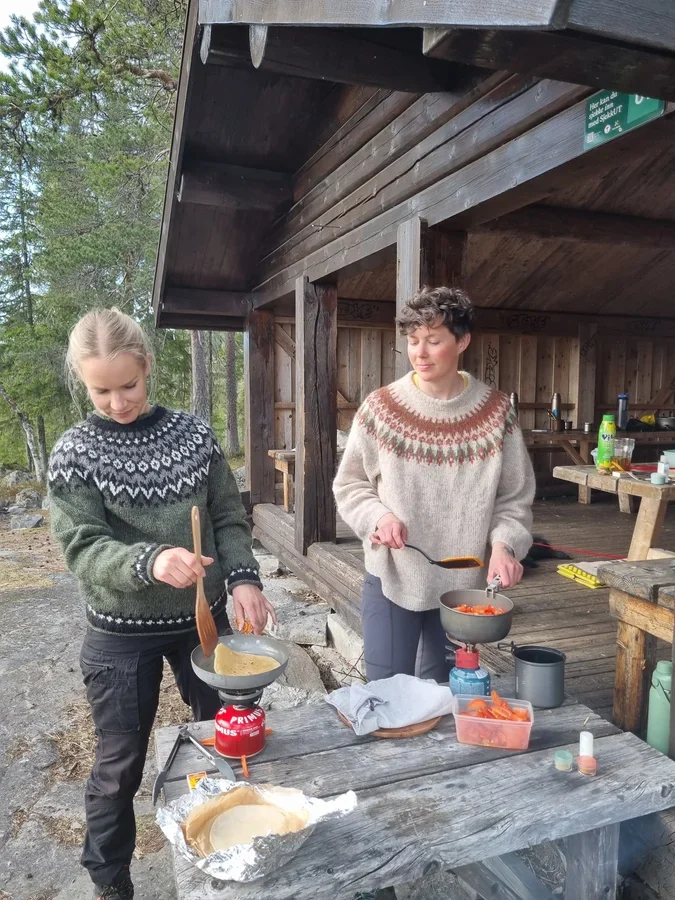 Two women standing in front of a wooden shelter in the forest cooking food on gas stoves