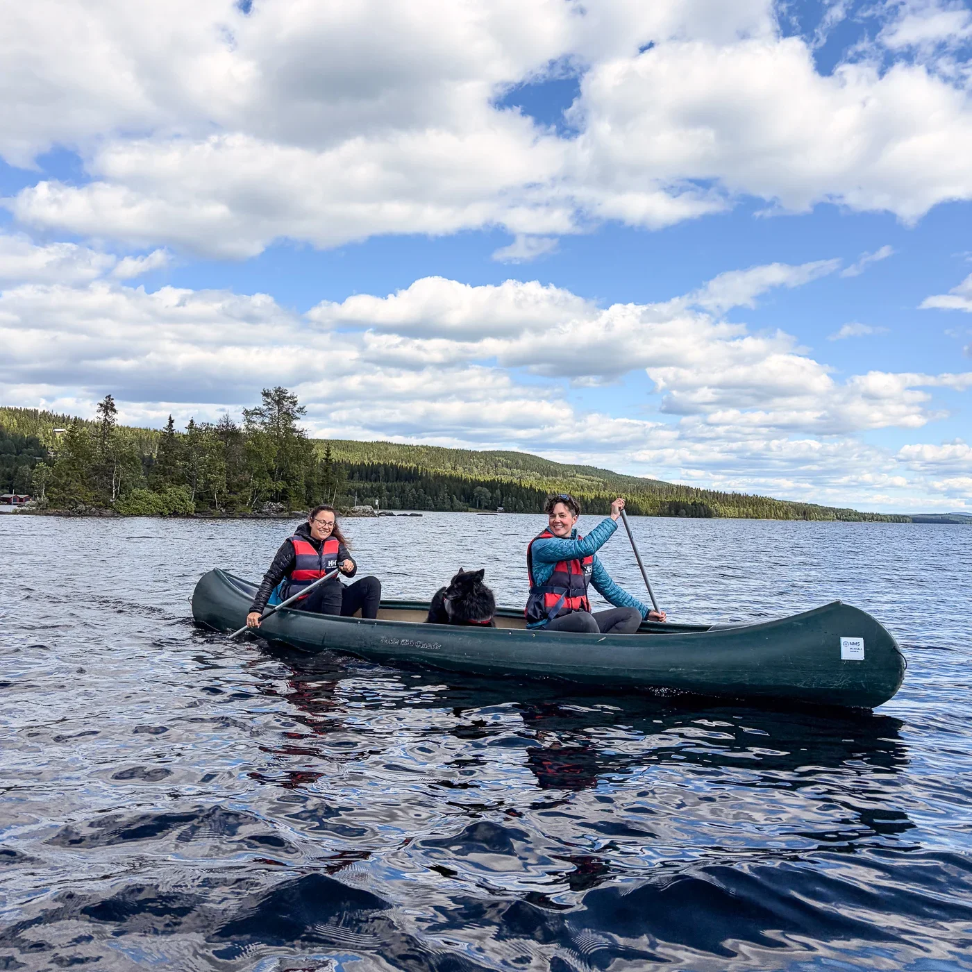 Two women and a black dog sitting in a green canoe and paddling on a big lake. In the distance the shoreline with trees and gentle hills is visible.