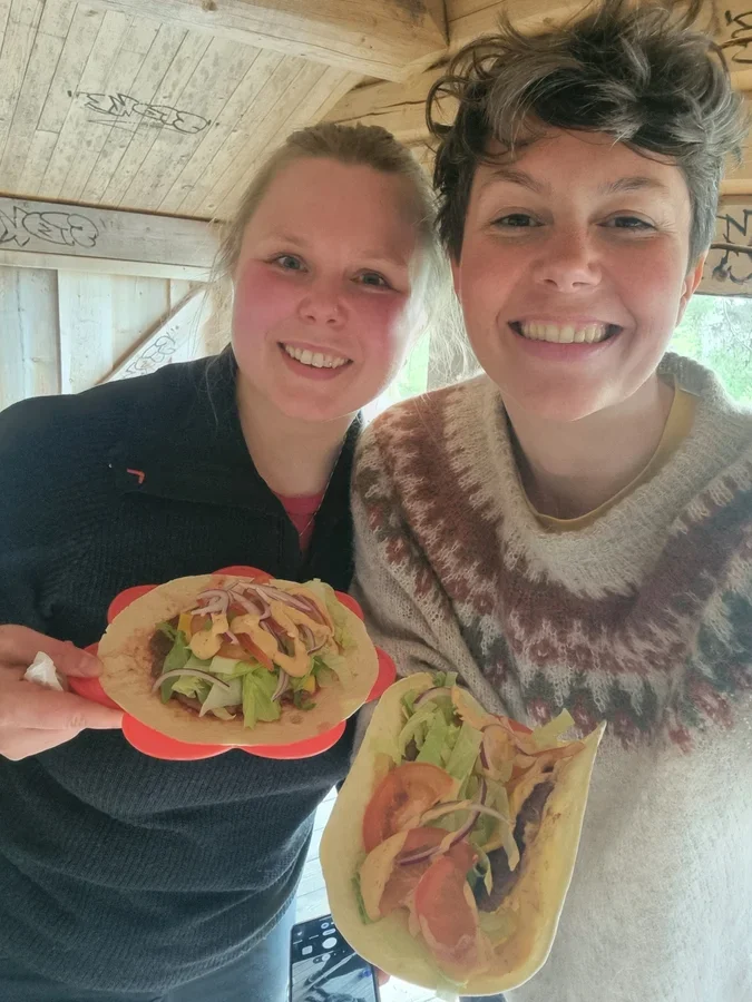 A selfie of two women in woolen sweaters holding food and smiling widely into the camera