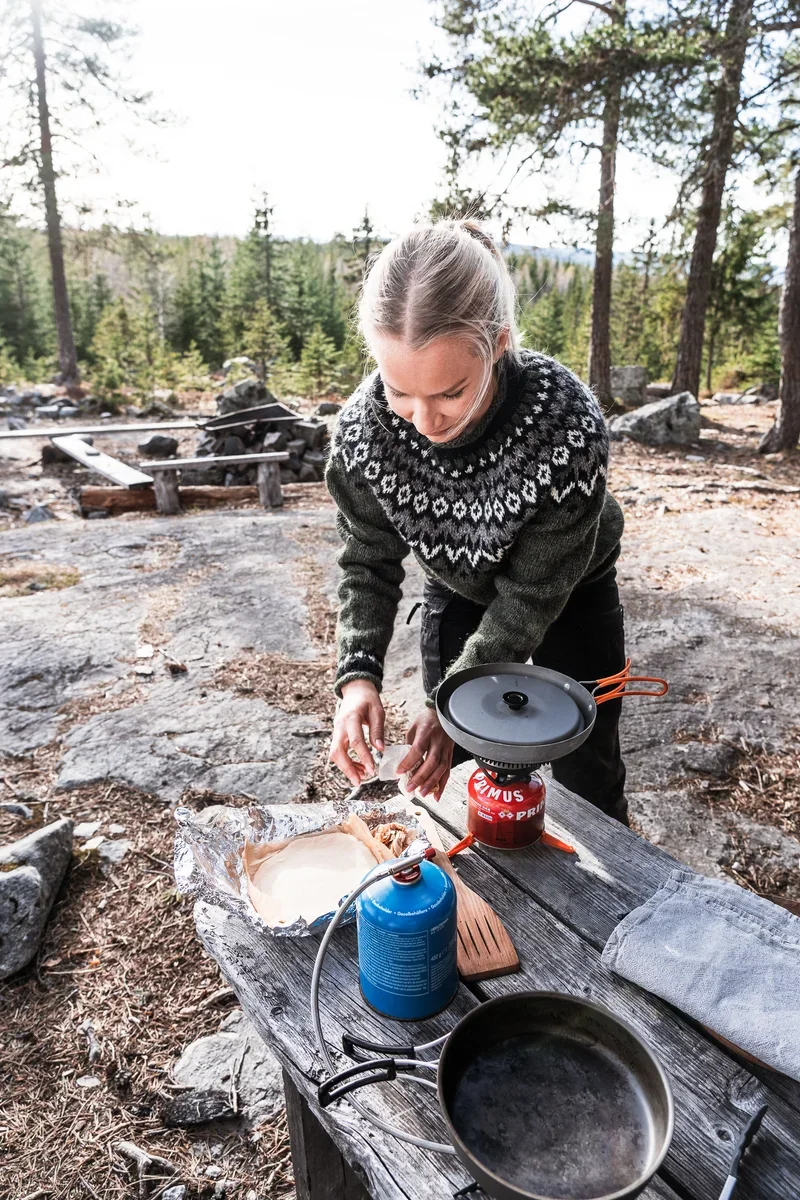 A photo of a woman in a woolen sweater cooking on a gas stove on a wooden table. In the background you can see a firepit and pine trees.