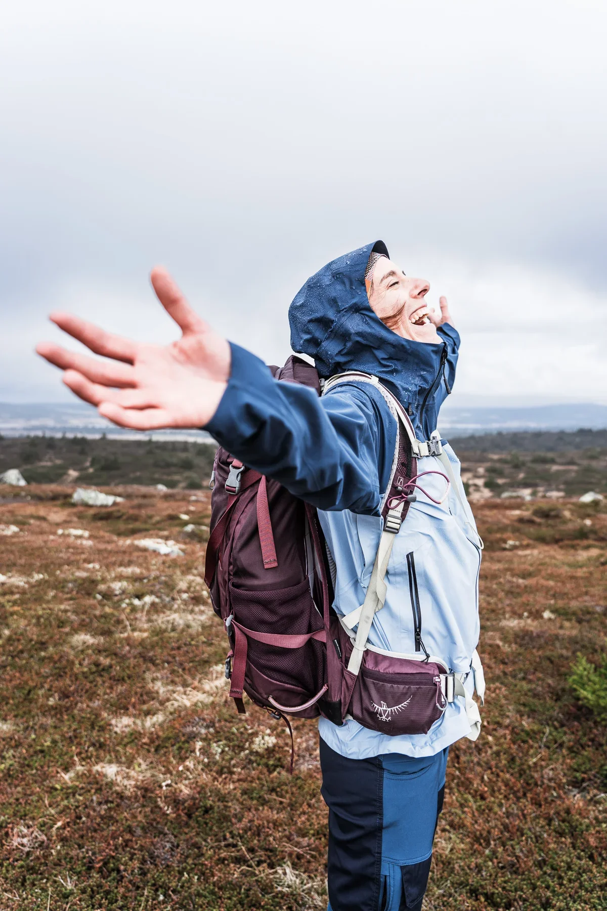 A woman with a blue rain jacket and a purple hiking backpack stretching their arms out and raising her head laughing. In the background you can see a hilly mountain landscape and grey clouds