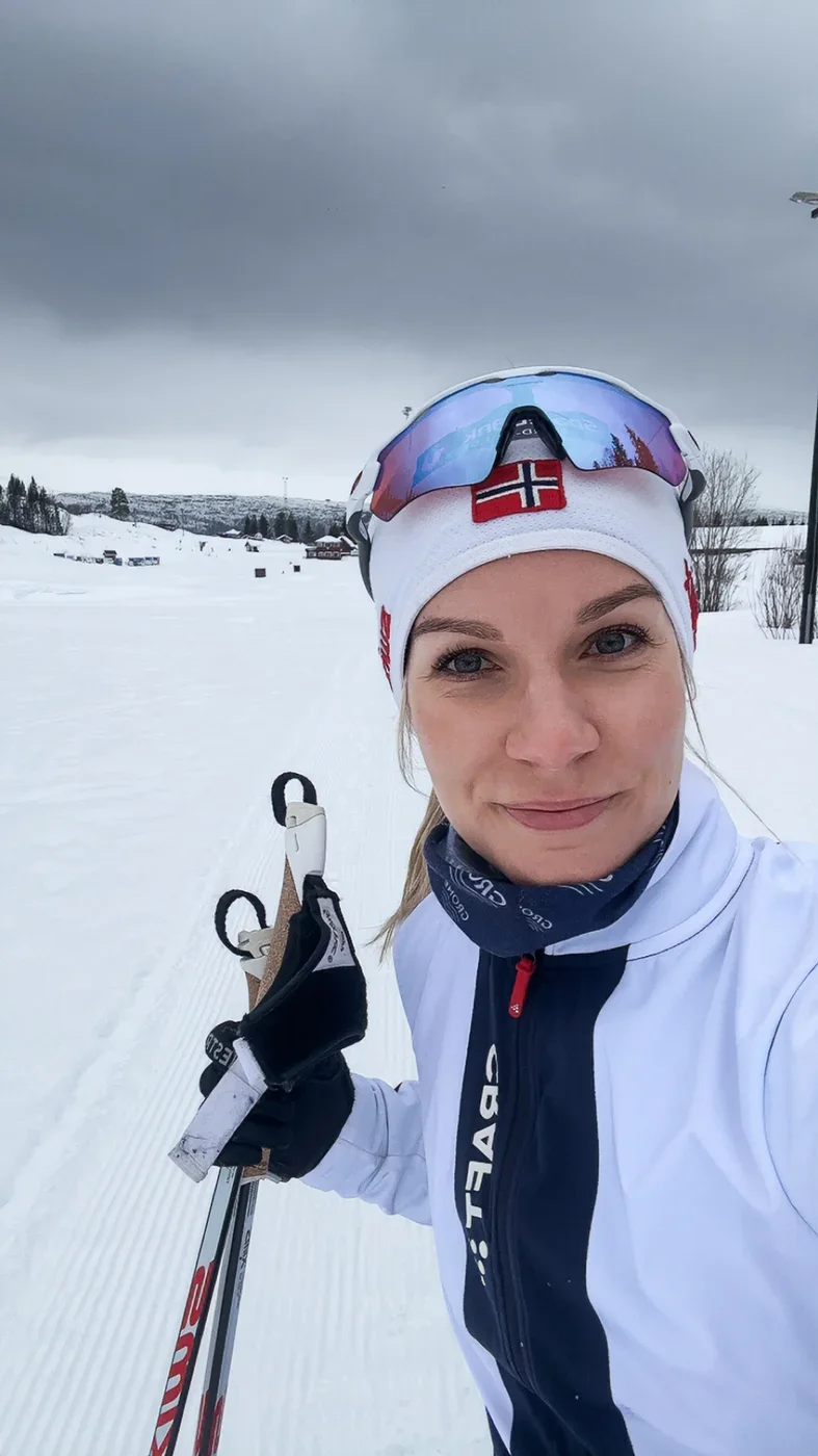 A selfie of a blonde woman in a white-blue cross country skiing jacket, a hat and glasses, smiling into the camera, holding skiing poles