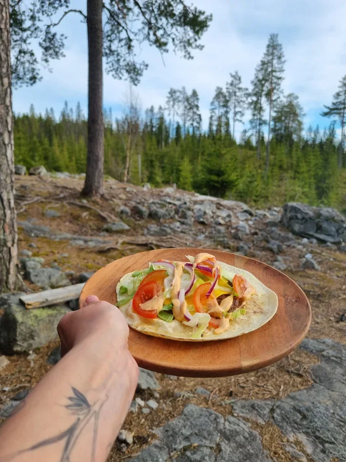 A photo of a hand holding a plate with a a wrap with tomato, onions and lettuce in front of blurry pine trees in the background