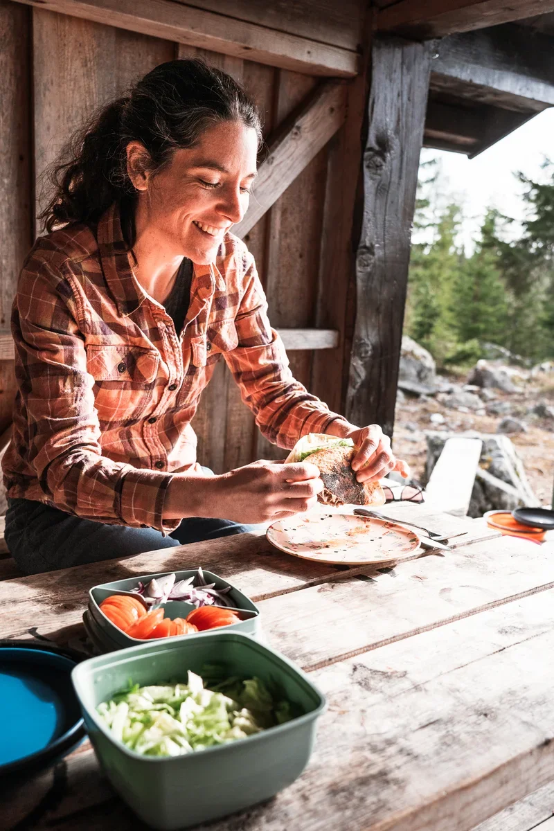 a photo of a woman in a lumberjack shirt sitting in a wooden shelter at a table eating a wrap. In front of her on the table you can see boxes with cut tomato and lettuce.