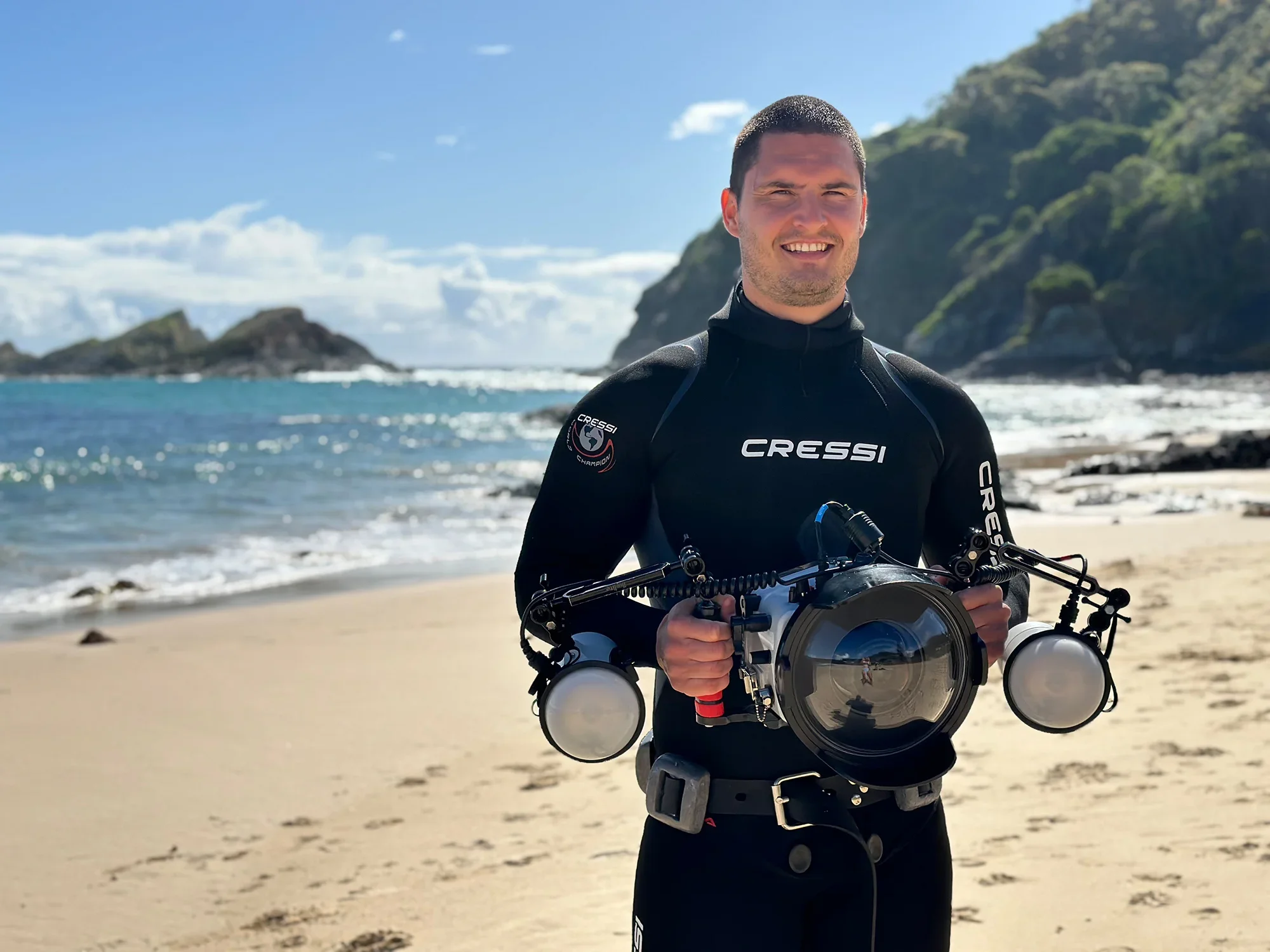 Man in wetsuit holding underwater camera gear on beach with ocean, rocks, and hills in background