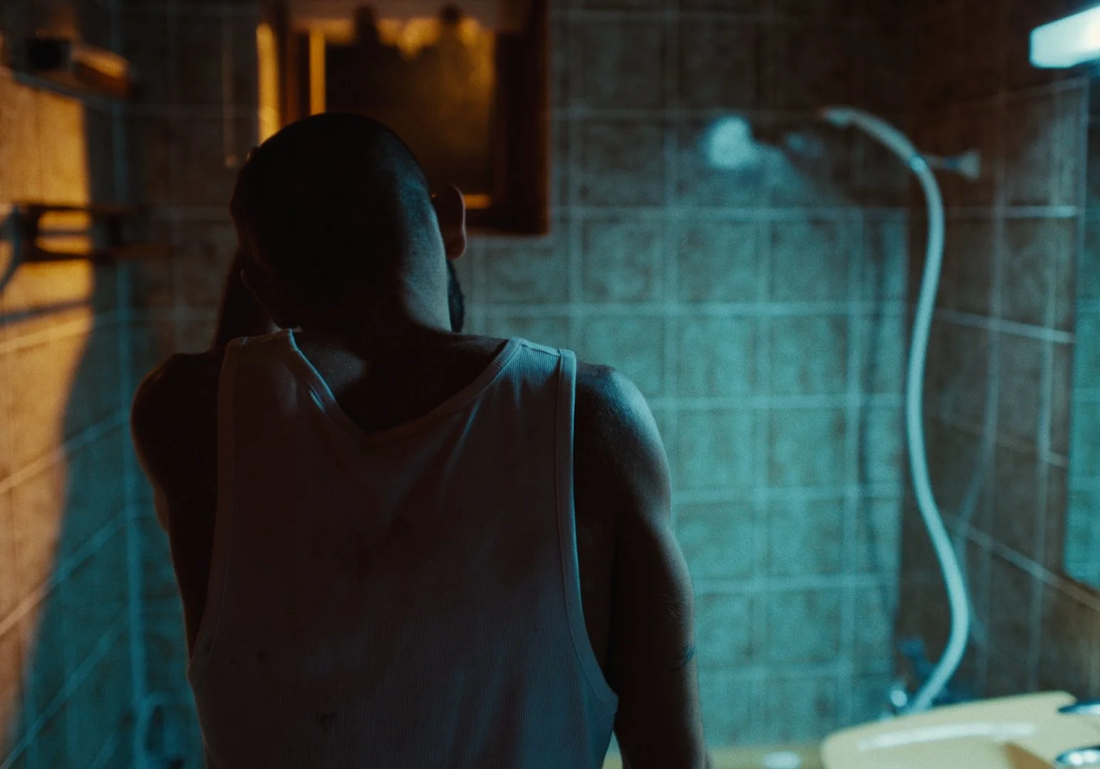A man with a beard wearing a white tank top, viewed from behind, standing in a dimly lit bathroom with brown tiled walls and a shower with a flexible hose.