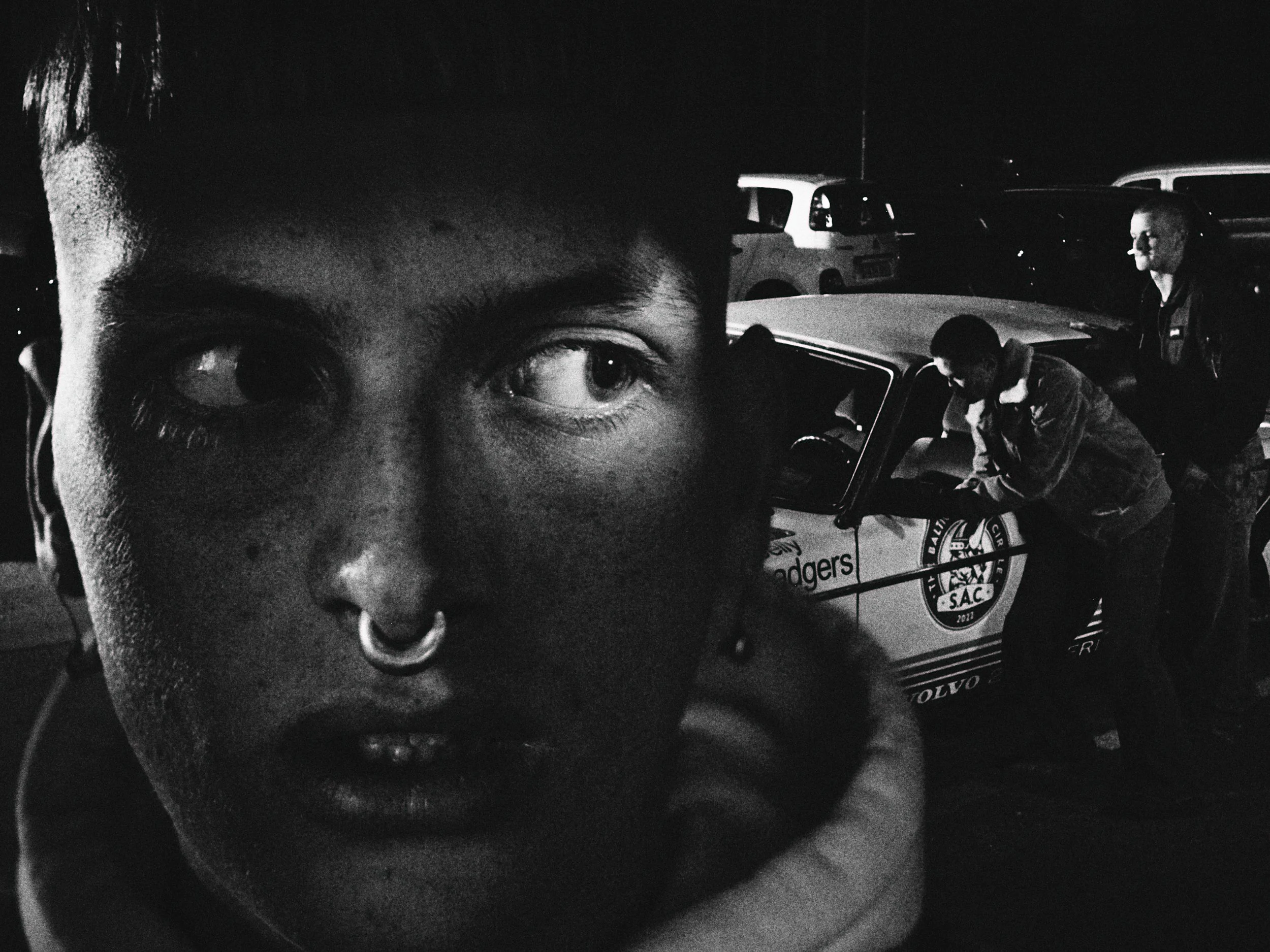 A close-up black and white photograph of a person's face with a septum piercing and an intense gaze, overlapping a background scene of two other people next to a police car, with parked cars in the distance.