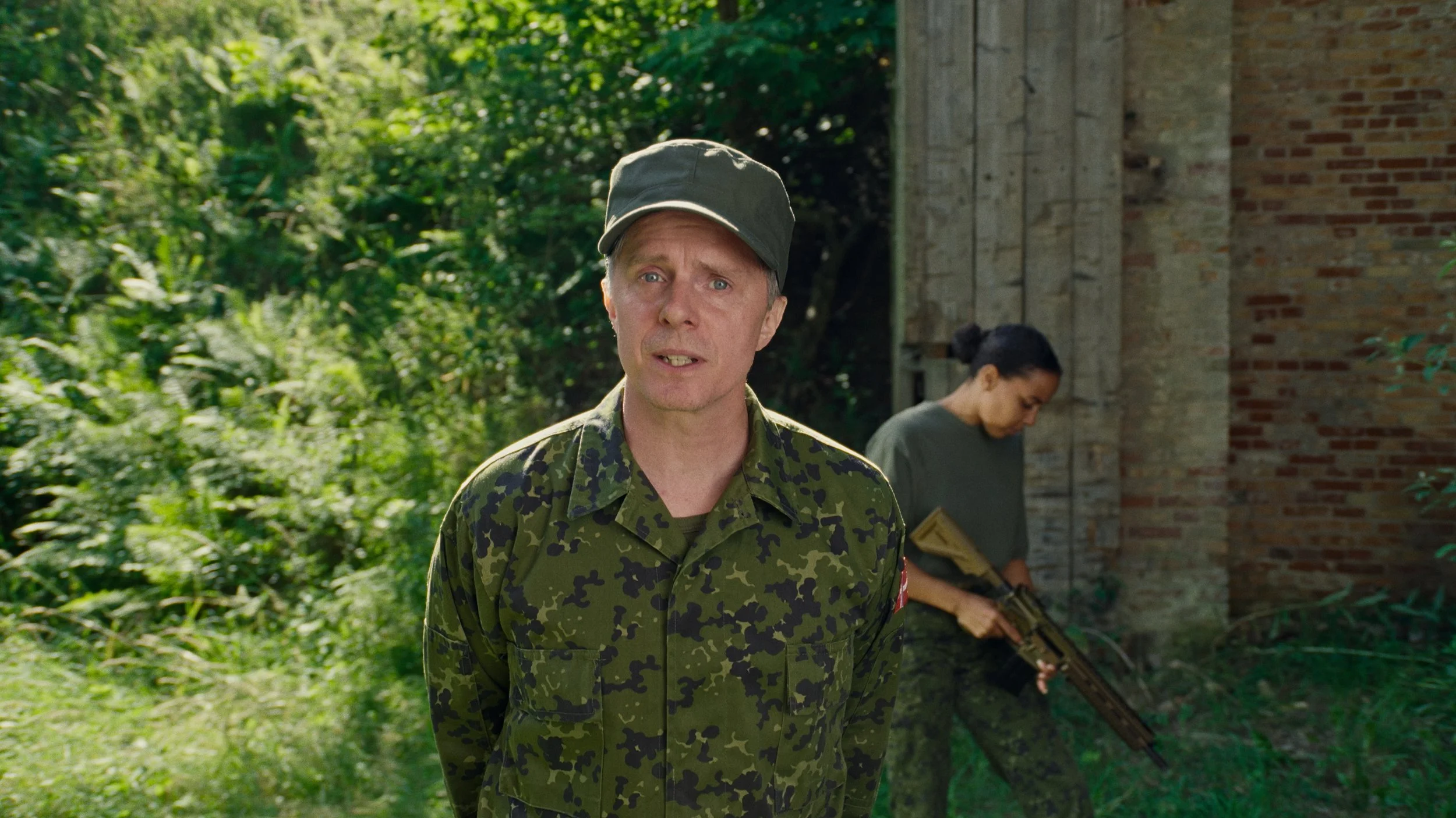 A man in a camouflage military uniform with a green cap standing outdoors in a lush, green forest, with a woman holding a firearm in the background near a brick wall.