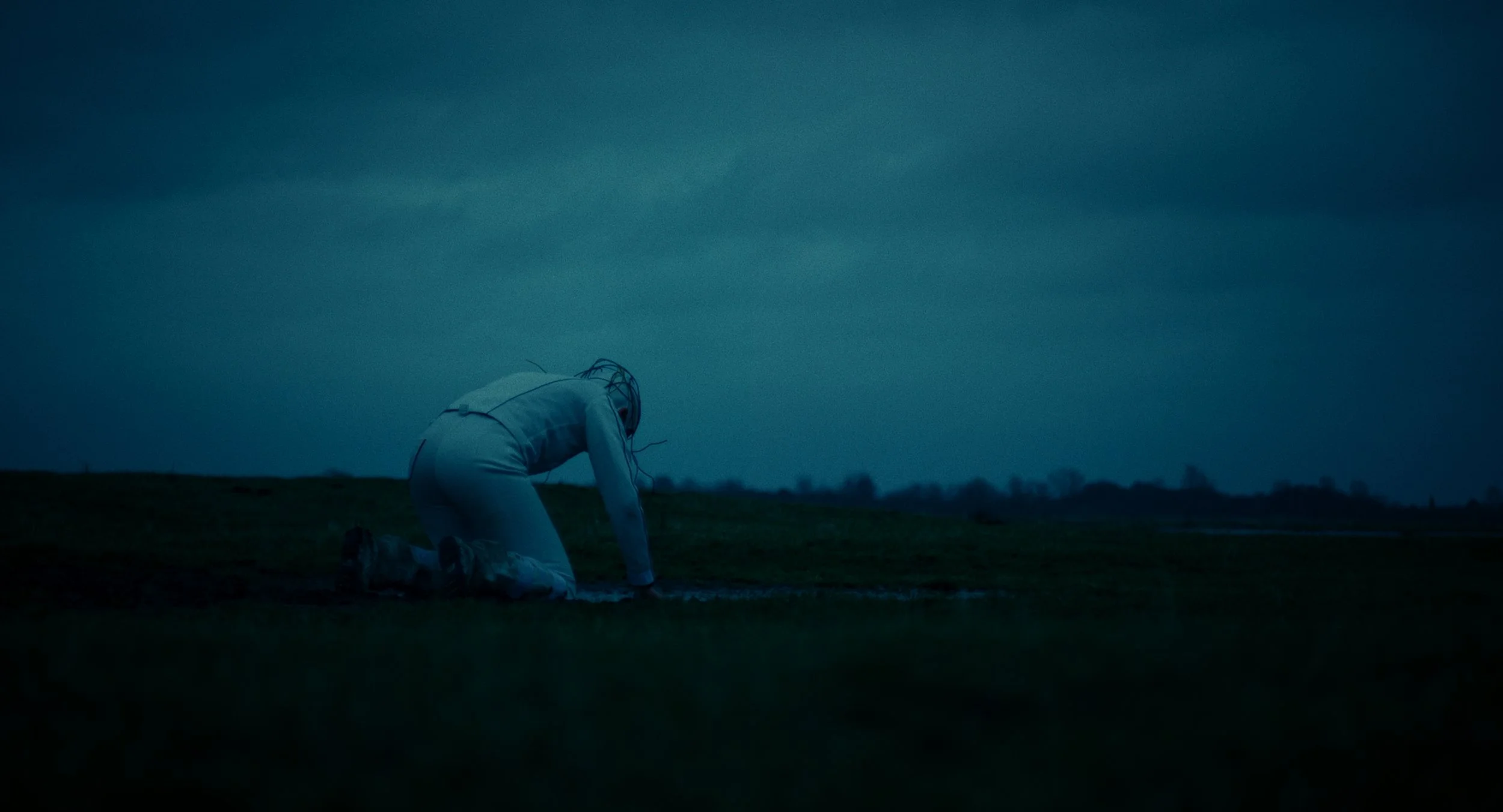 A person kneeling on the ground outdoors in a dark, cloudy setting, with their head bowed and hands on the ground, wearing light-colored clothing.