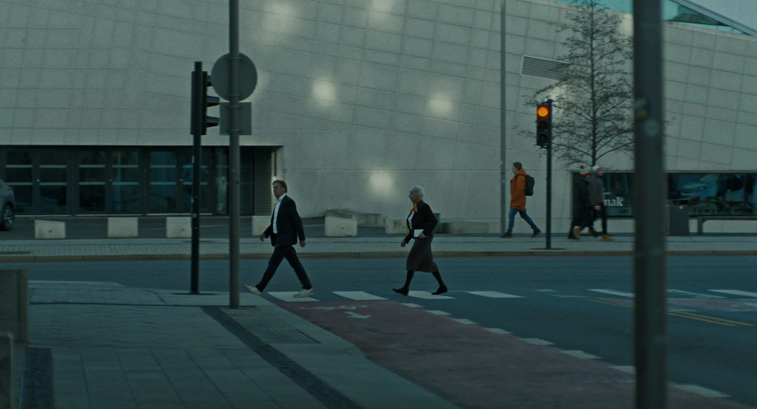 People crossing a city street at a crosswalk with a traffic light showing red, in front of a modern building.