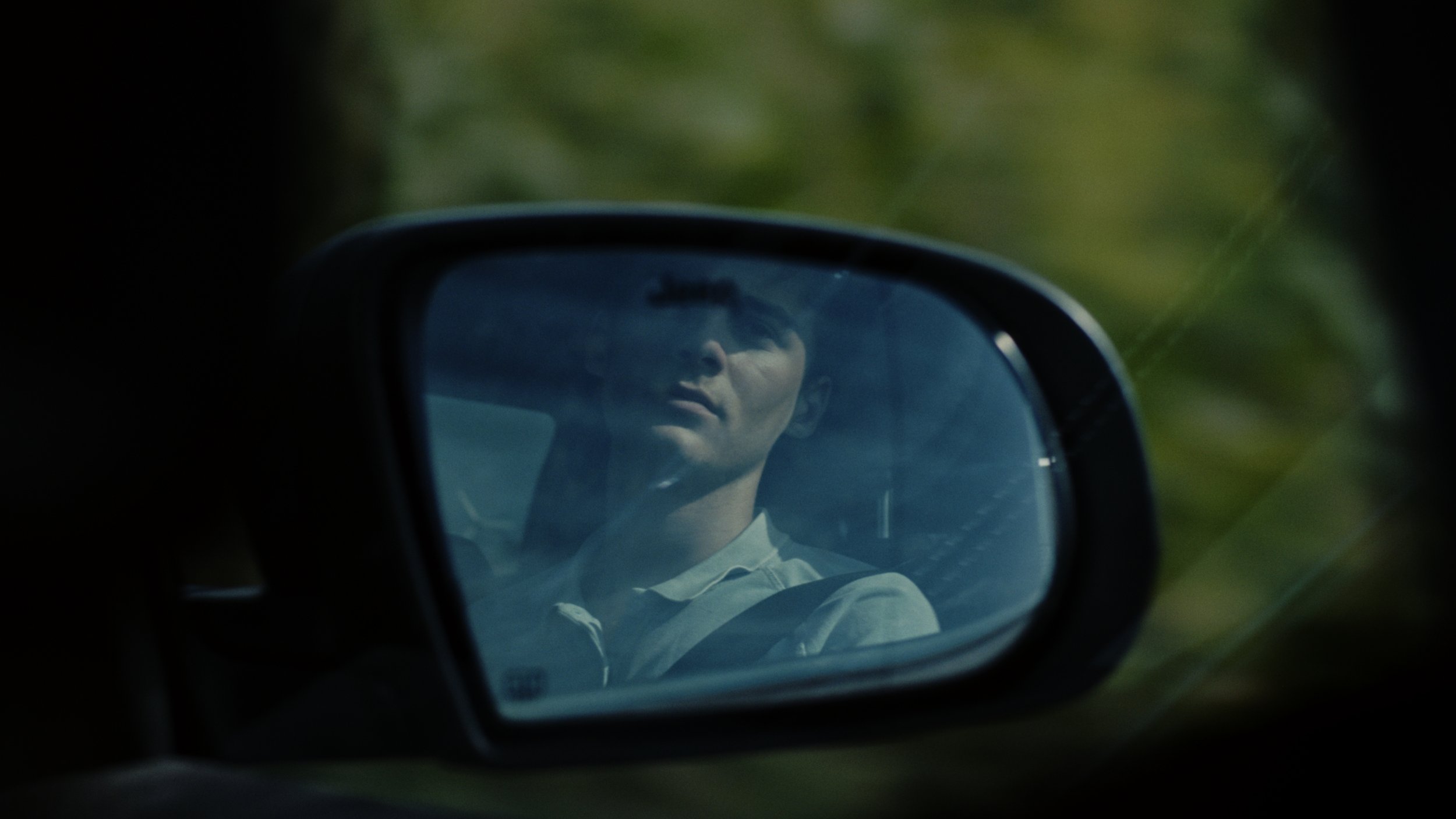 A man looking at himself in a car's side mirror, with green foliage in the background.