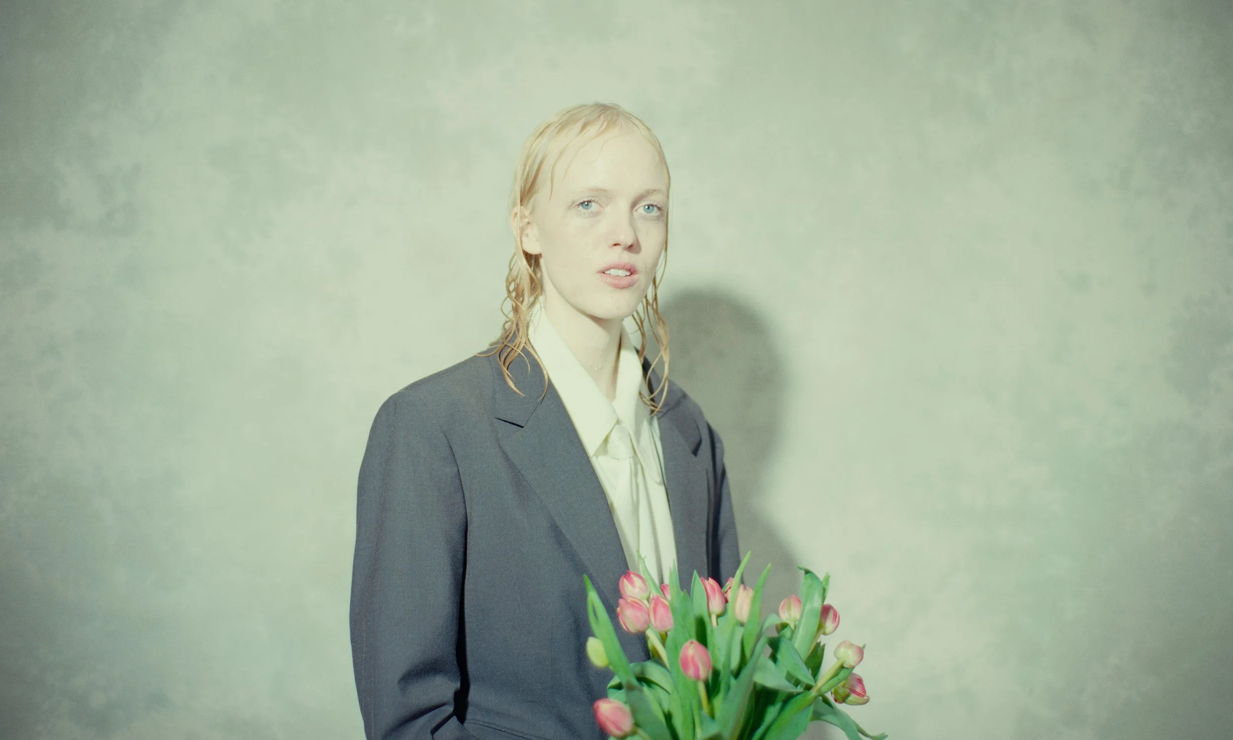 Woman with wet hair and a serious expression holding a bouquet of pink tulips, wearing a gray blazer and cream blouse, against a plain light-colored background.