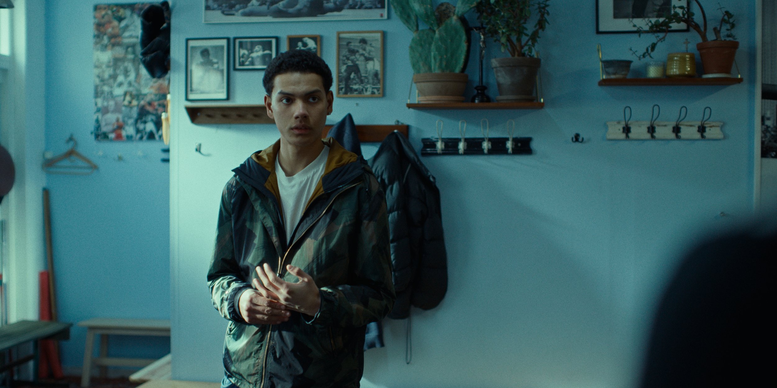 A young man with short curly dark hair wearing a camouflage jacket and white T-shirt standing in a room with light blue walls, decorated with framed photos, potted plants, and hooks on the wall.
