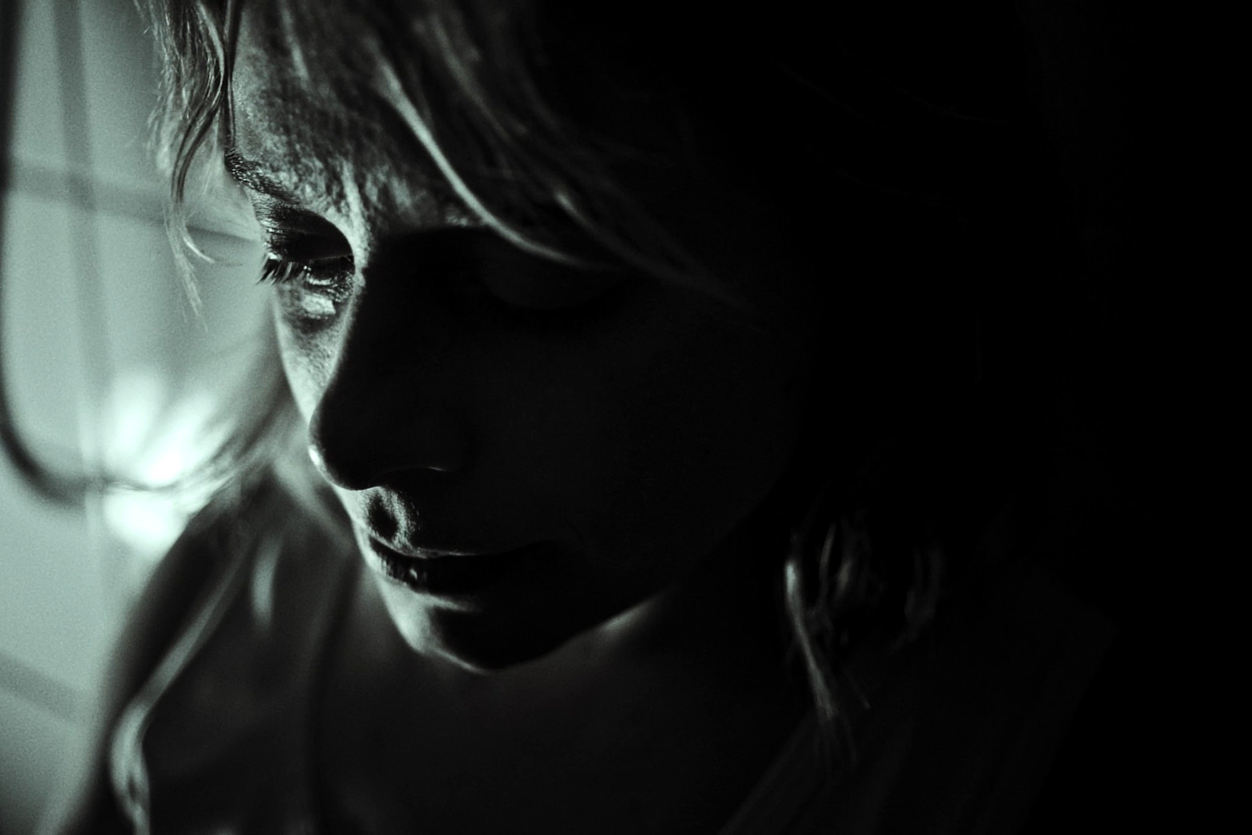 A black and white close-up of a woman's face, with shadows obscuring much of her features, showcasing her nose, closed eyes, and eyelashes, with her hair partially visible.