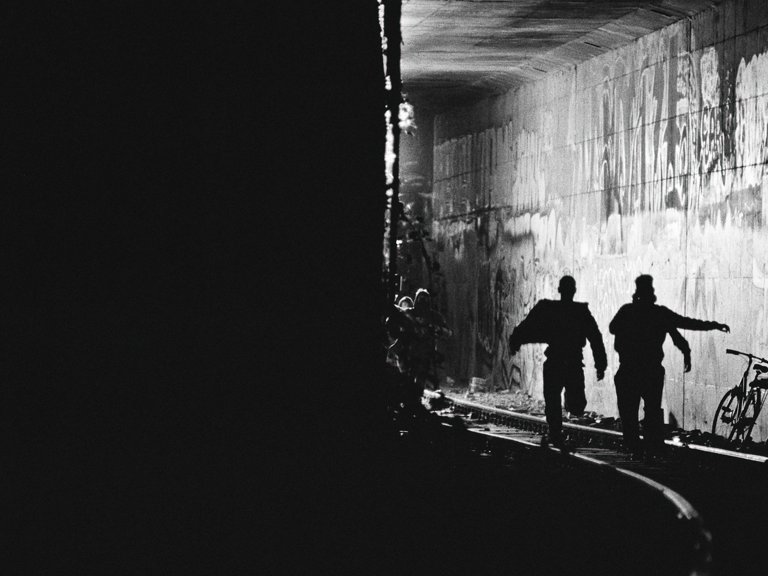 Silhouettes of two people walking in a graffiti-covered tunnel, with bicycles leaning against the wall, in black and white.