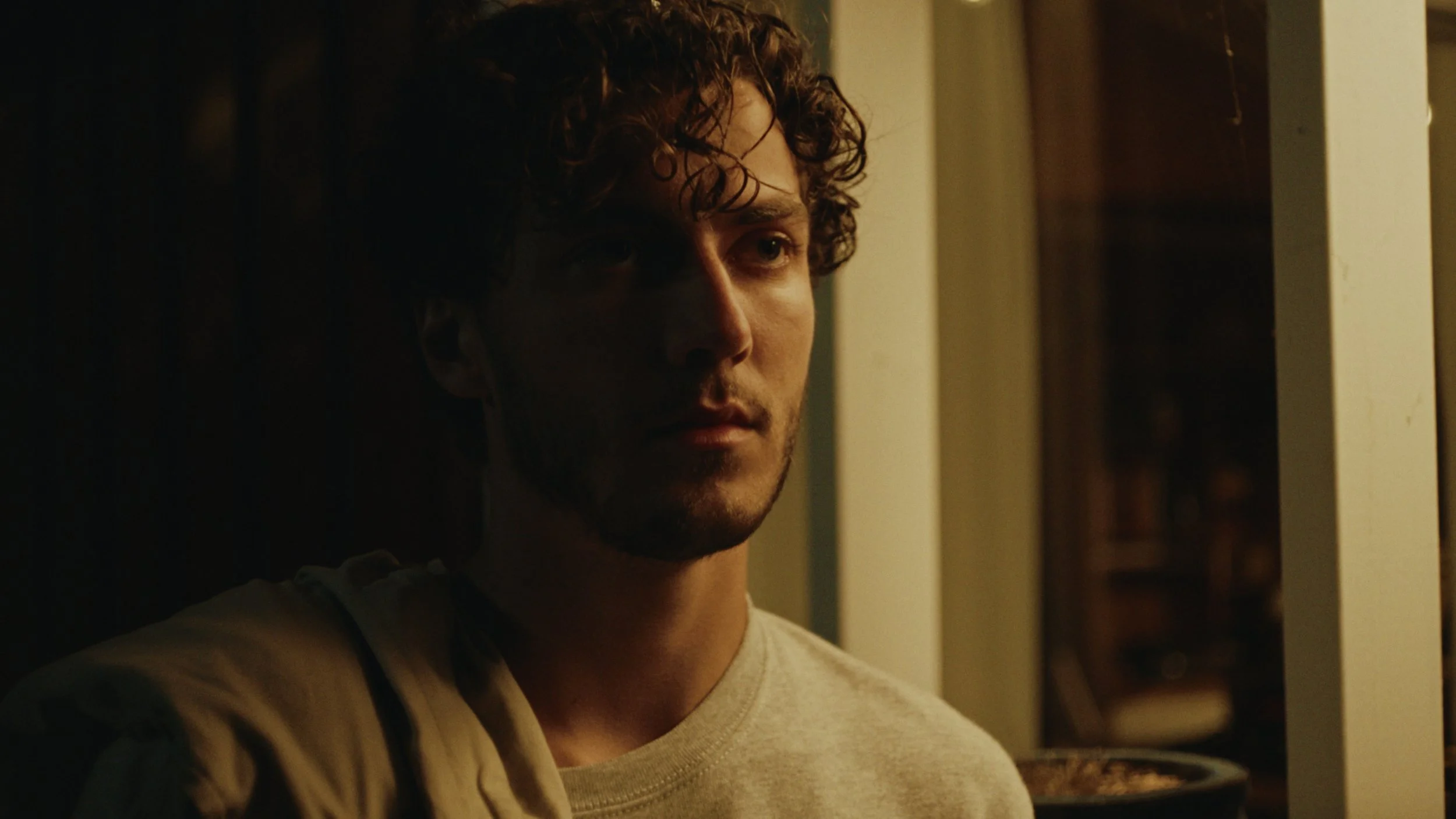 A young man with curly hair looks pensively out a window in low lighting.