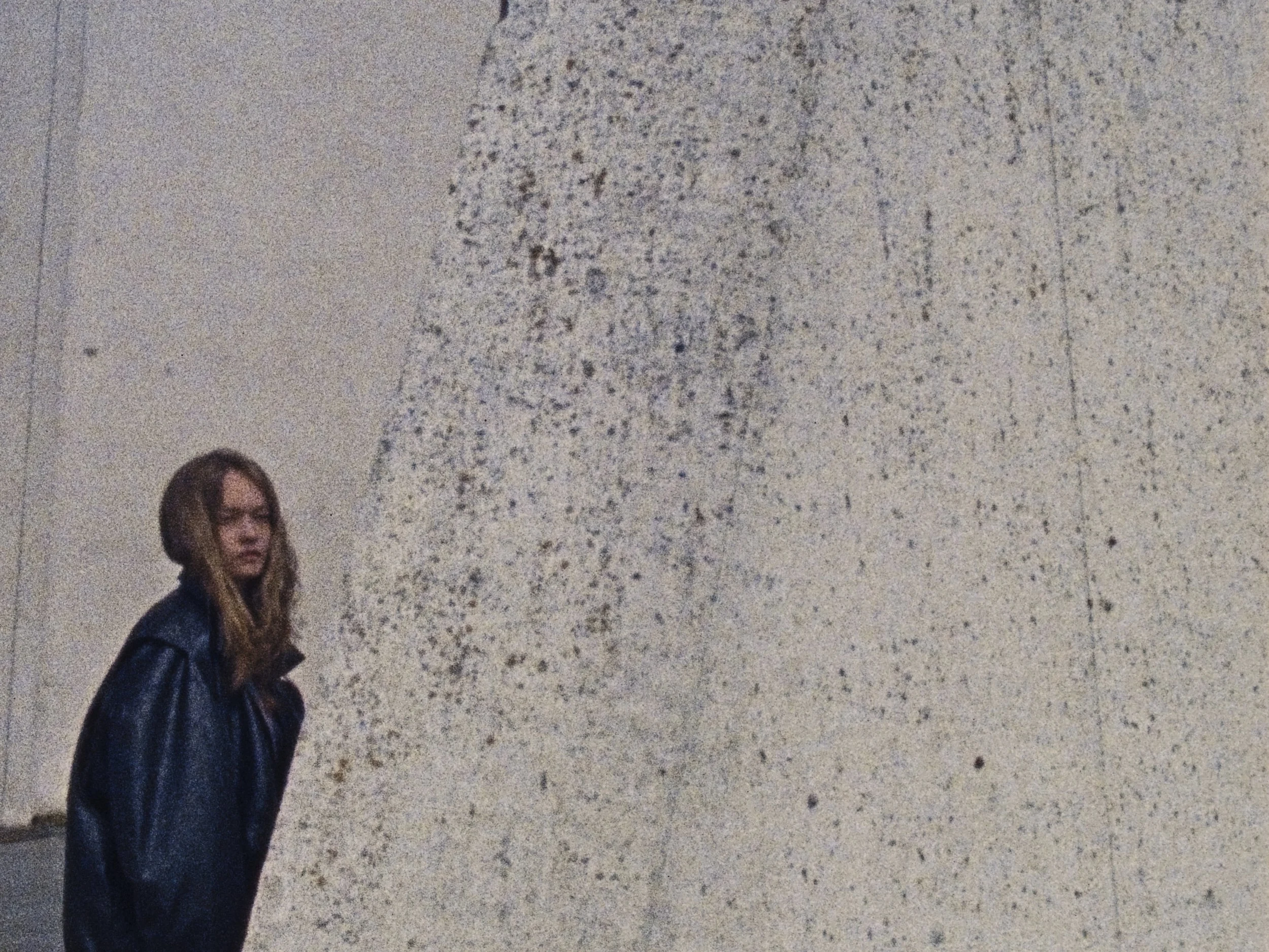 A woman with long hair wearing a black leather jacket stands next to a large, textured stone wall.