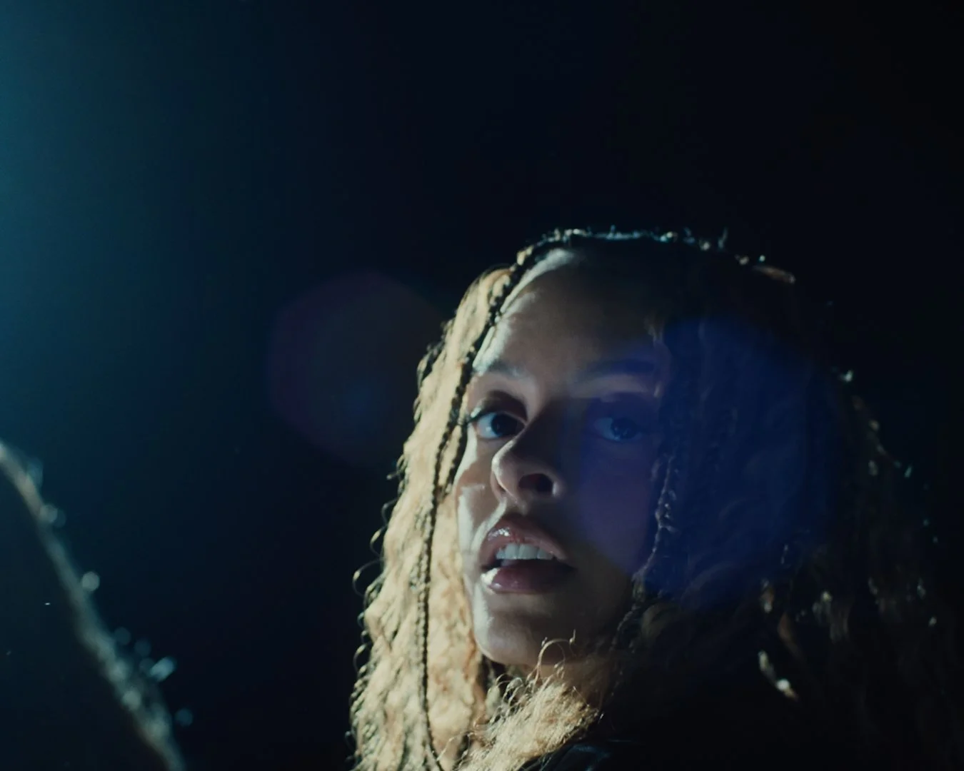 Close-up of a woman with styled braids, looking upward in a dark setting with blue lighting