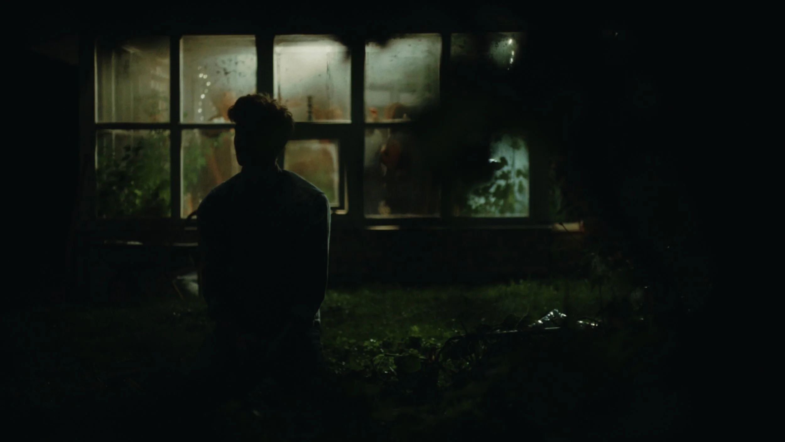 A person kneeling outside a house at night, with a window in the background showing faint indoor light and reflections, surrounded by darkness and some foliage.