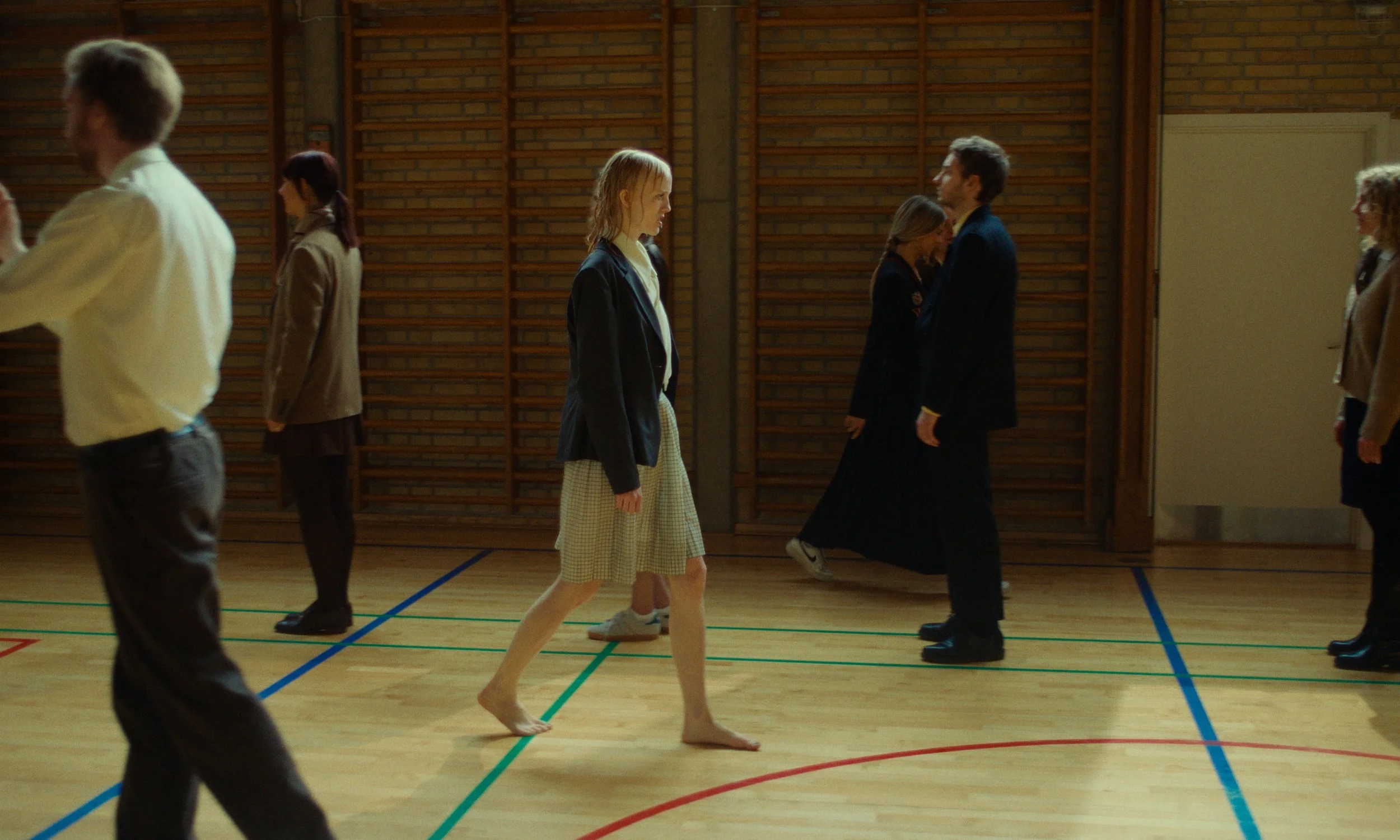 Multiple people standing and walking in a gymnasium or sports hall with wooden lining on the walls, some dressed casually and others in dark attire, and one woman walking barefoot on the wooden floor.