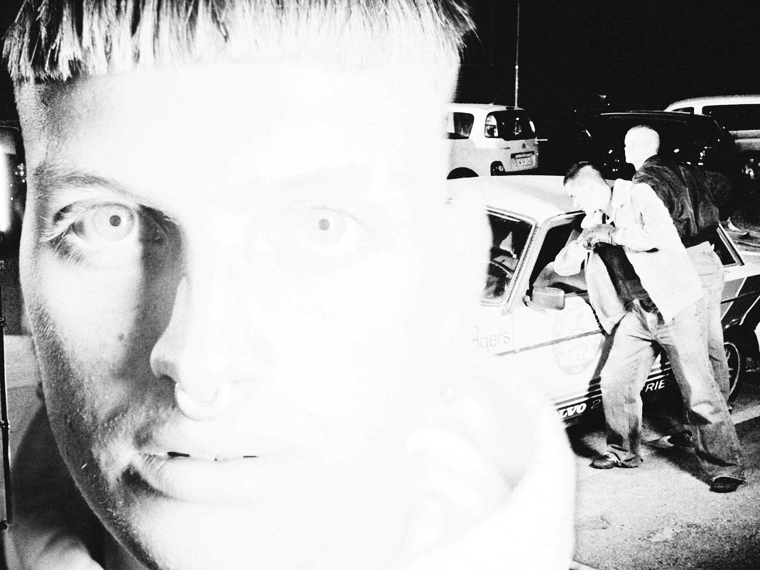 Black and white photo of two men clashing with a car in a parking lot at night, with a large blurred face of a person in the foreground.