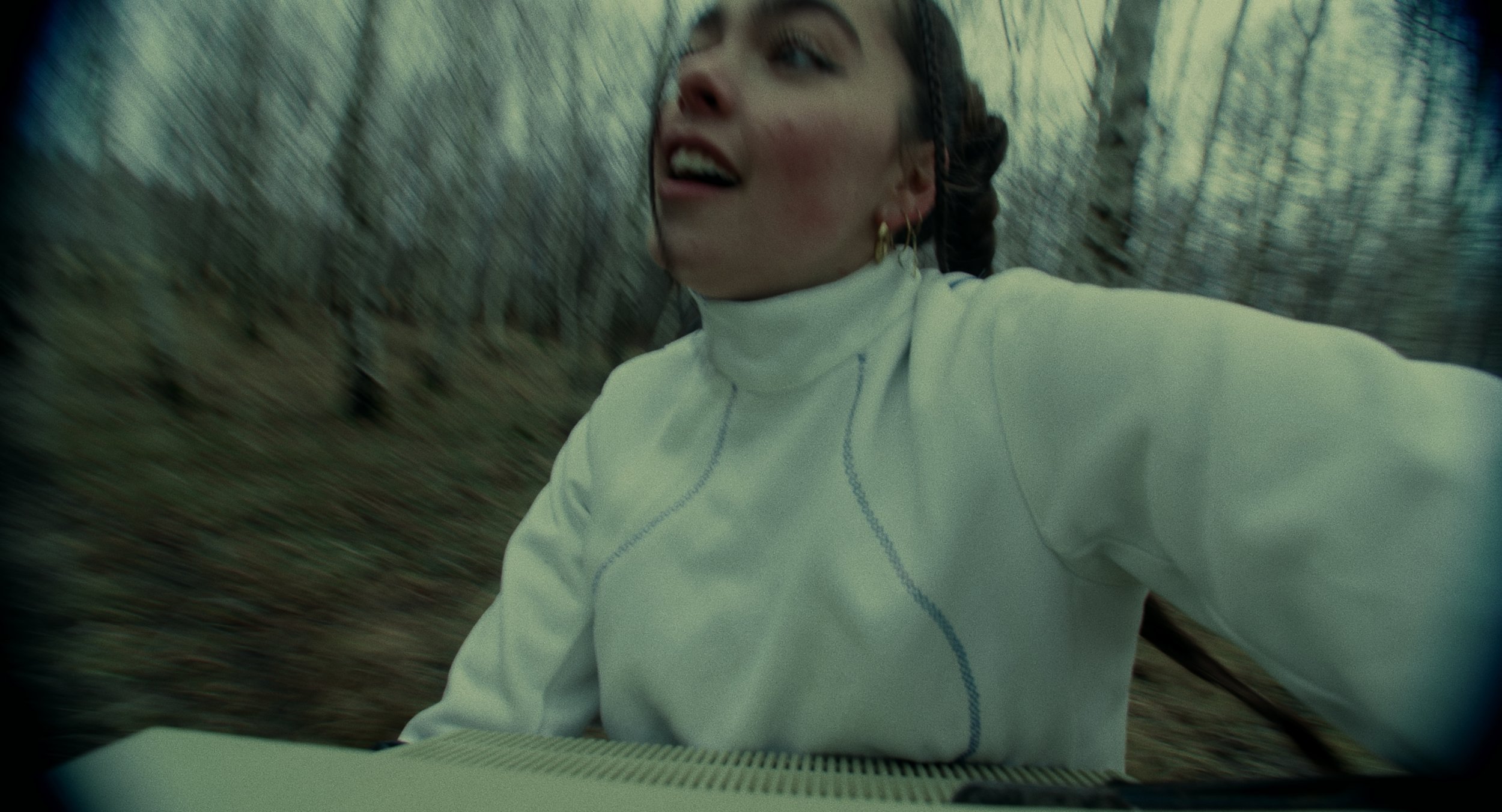 A woman in a white turtleneck top with blue stitching and gold earrings is riding a bicycle through a leafless forest, with a focused and slightly surprised expression, as the camera captures her from a low angle.